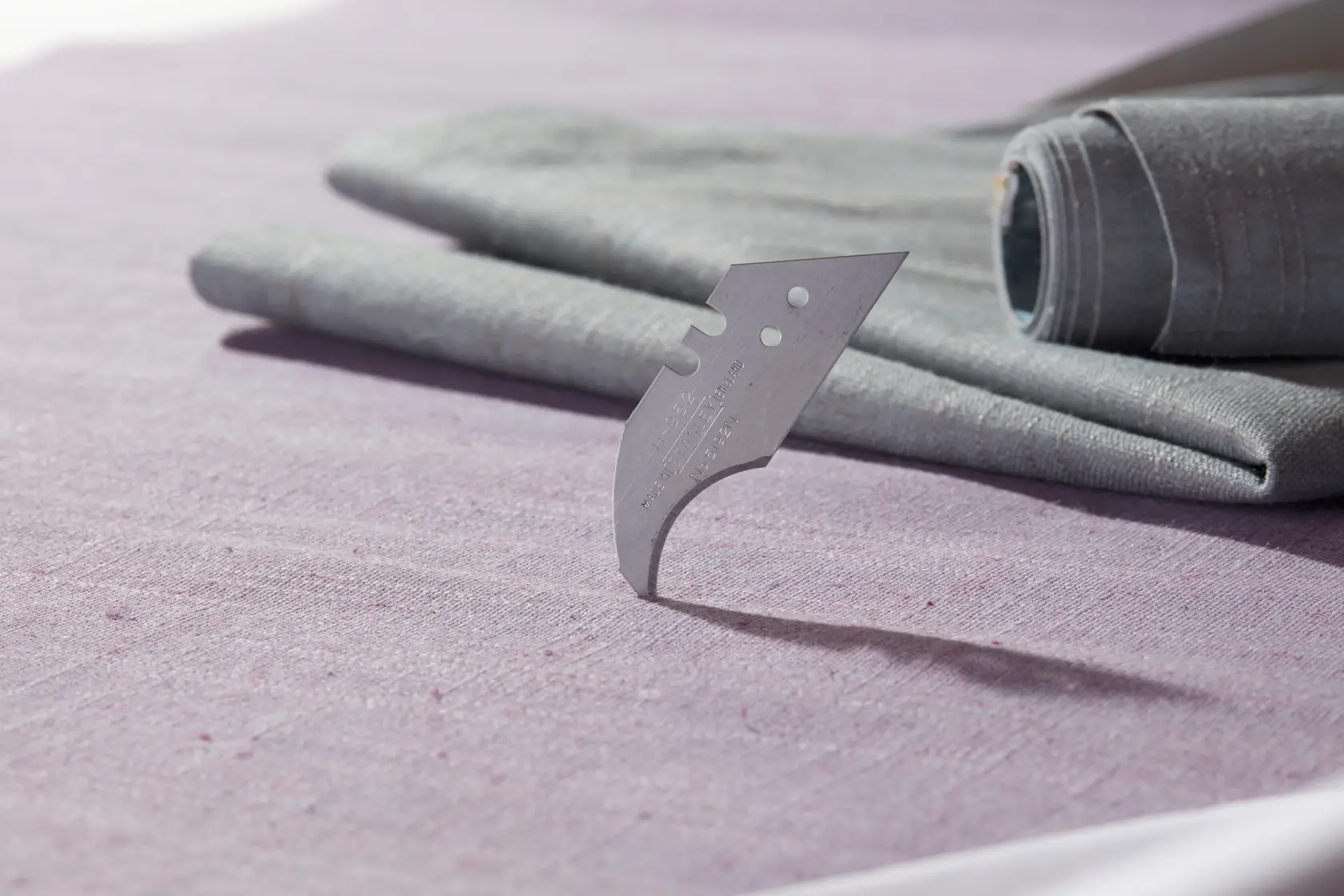 STANLEY 0-11-952 curved utility blade on purple fabric, two grey rolled textiles in background.