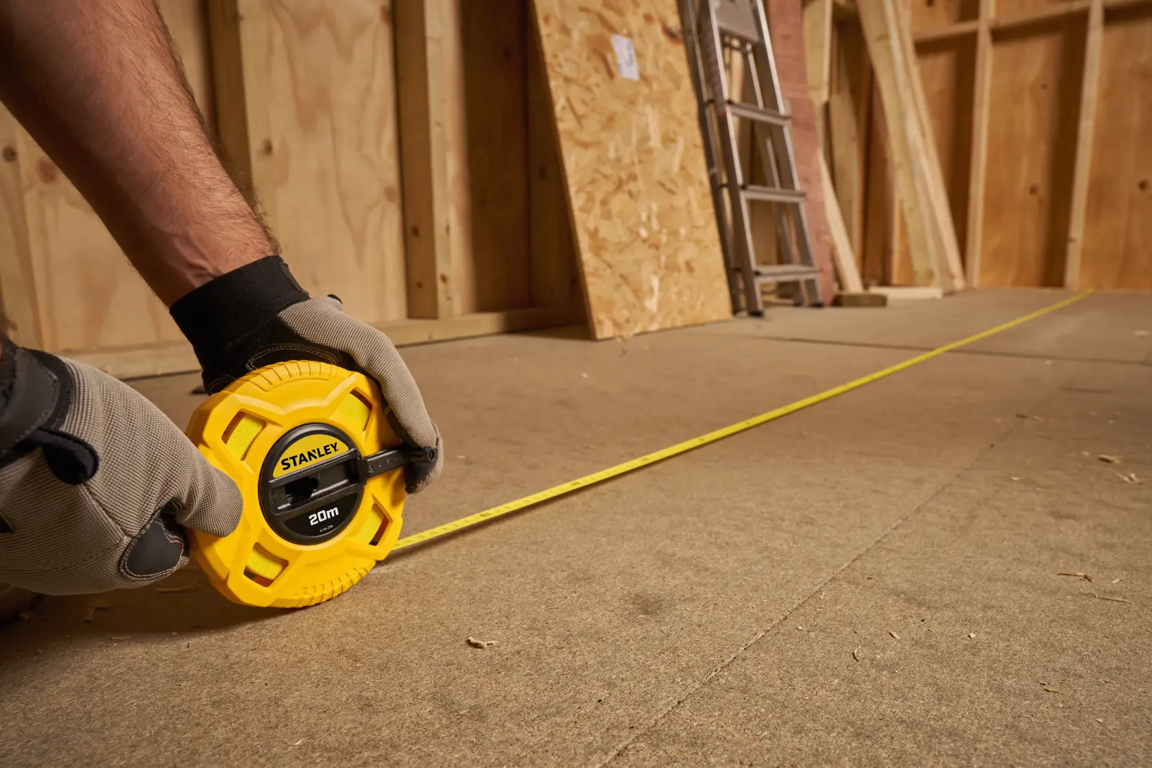 STANLEY 0-34-296 yellow tape measure, 20m, extended across a workshop floor, held by gloved hands.