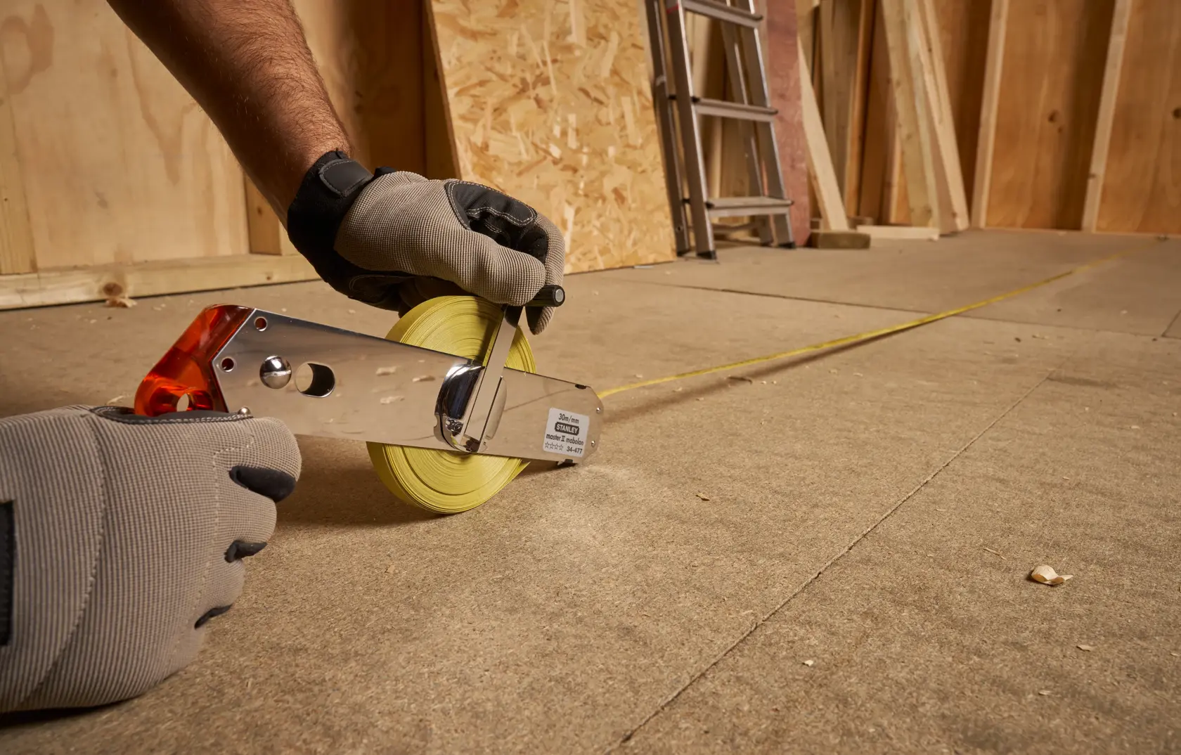 STANLEY 1-34-477 open reel tape measure with yellow blade, used by gloved hands on a construction floor.