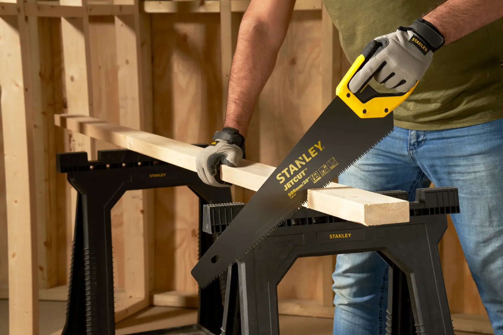 Photo of STANLEY 2-20-151 hand saw cutting wood plank, person wearing gloves, wood sawhorses in workshop.