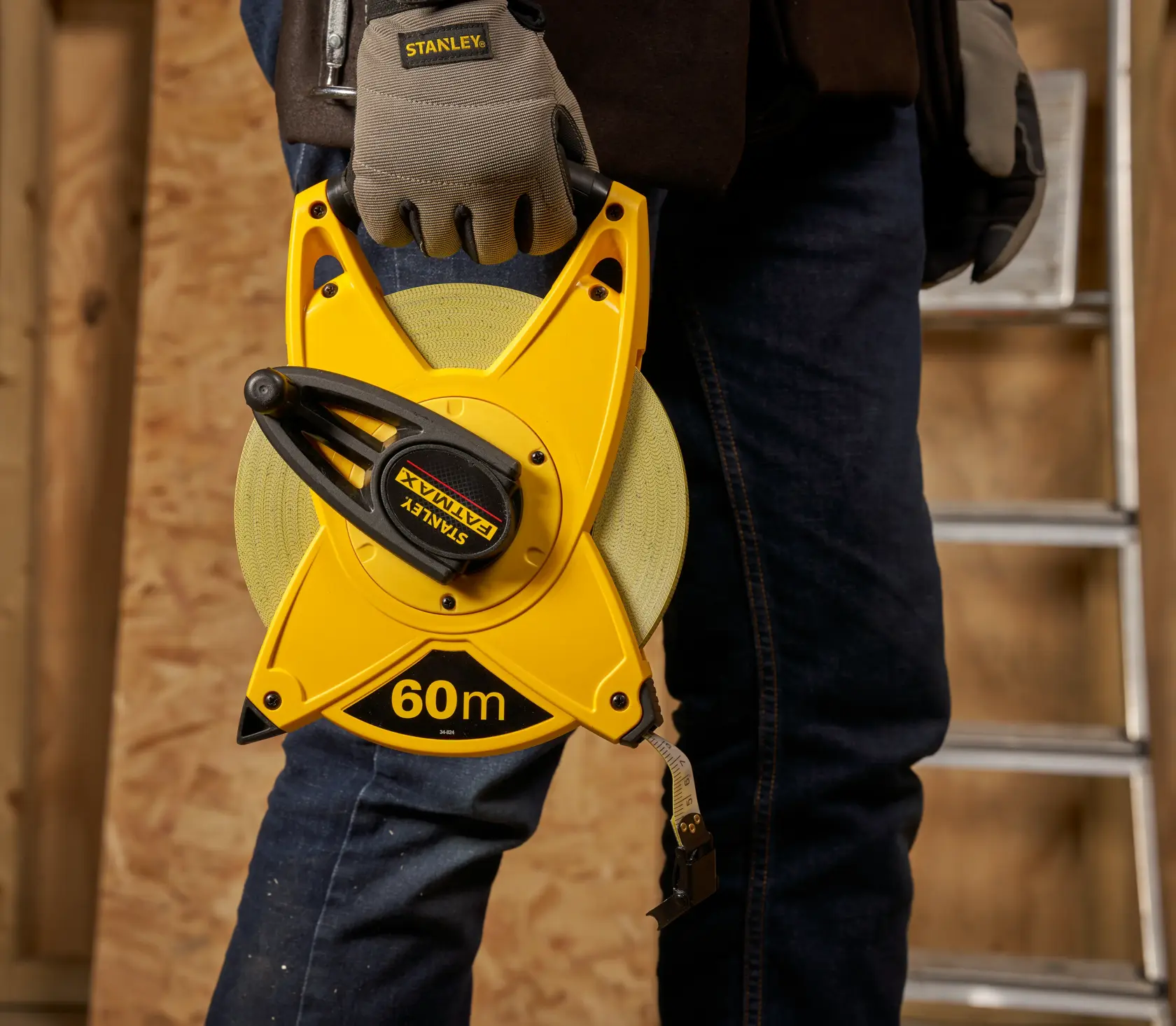 STANLEY 2-34-824 yellow 60m open reel tape measure held by worker in gloves, indoor workshop setting.