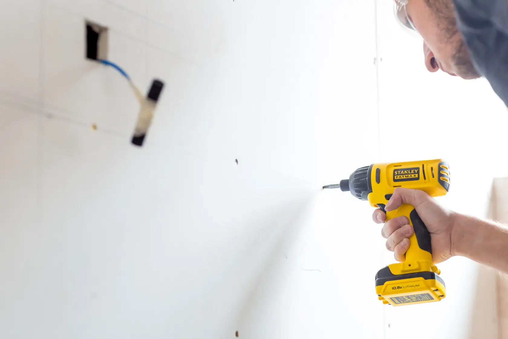Stanley FMC041S2 cordless drill in use, yellow color, shown drilling into white drywall.