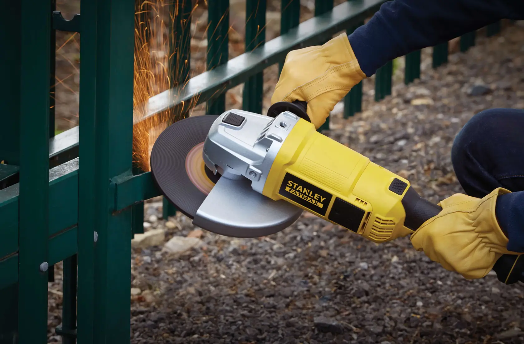 STANLEY FME841 angle grinder in use, yellow body, grinding metal fence, side view, sparks flying.