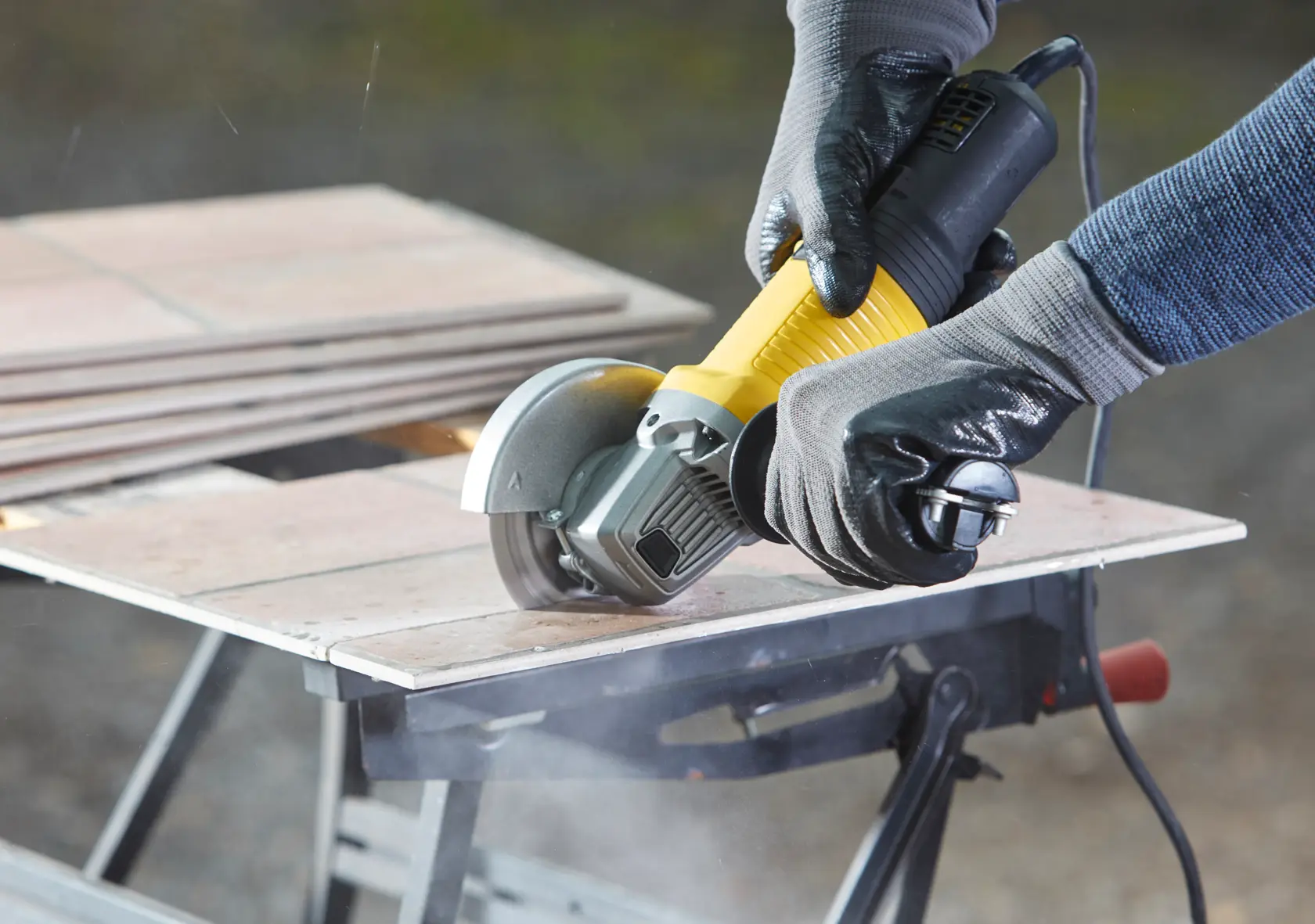STANLEY FMEG210K angle grinder cutting tile, close-up view, yellow and black tool, held by gloved hands.
