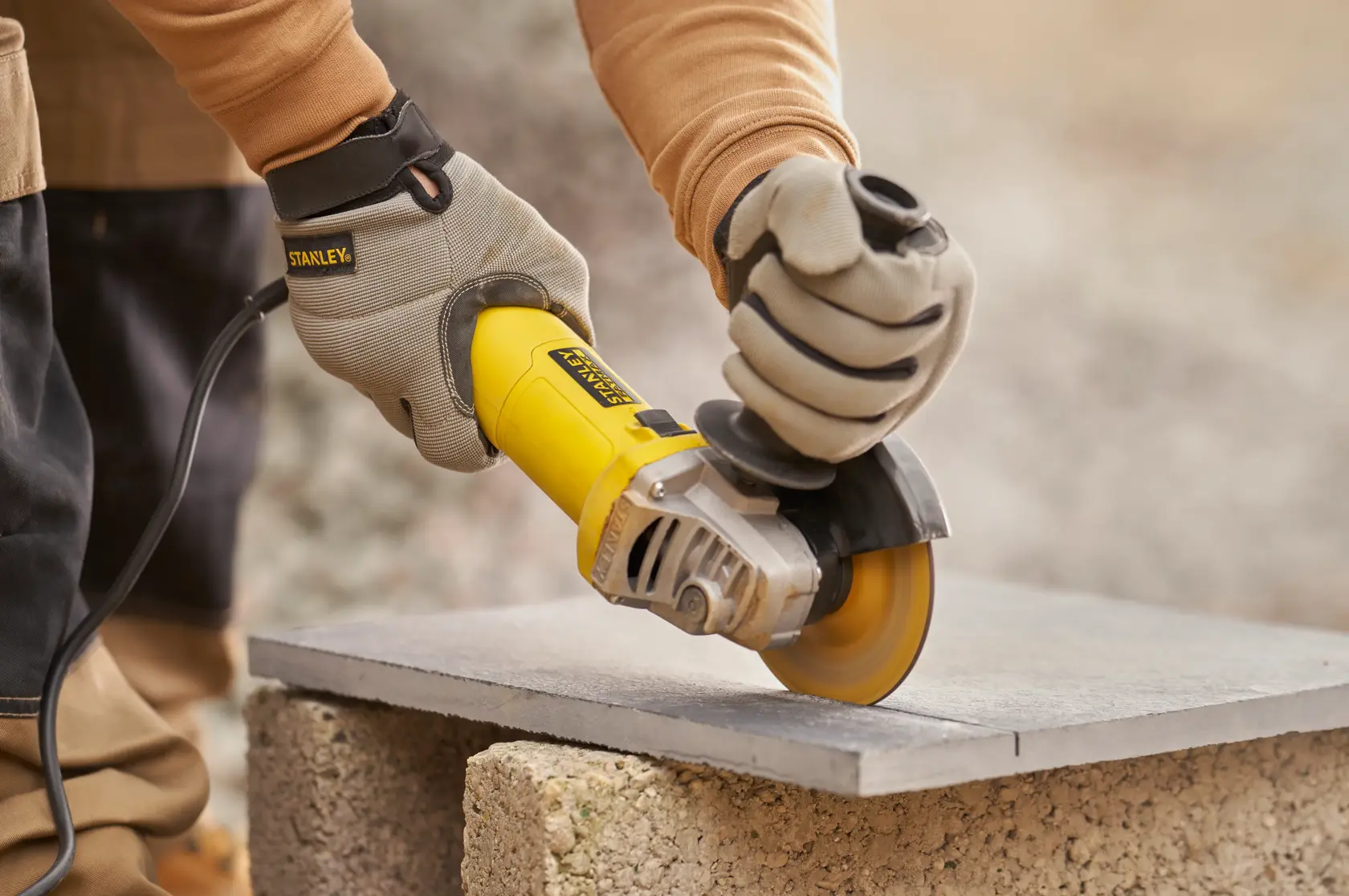Photo of STANLEY FMEG615 angle grinder in use cutting a gray tile, worker wearing protective gloves.