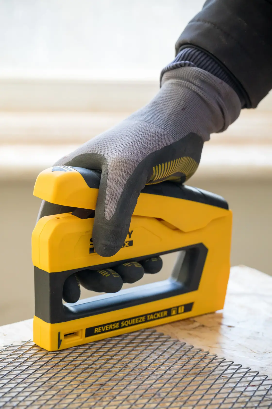 STANLEY FMHT0-80551 tacker in yellow and black, held by a gloved hand, pressing mesh onto wood surface.