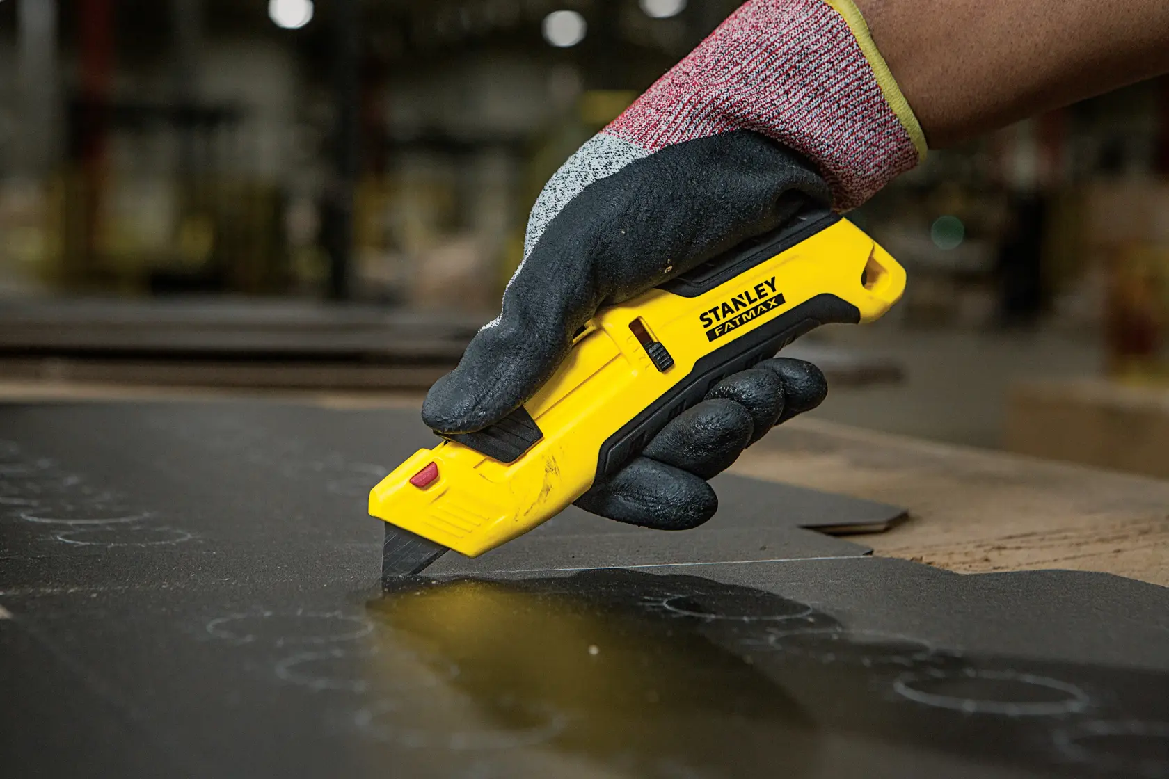 Photo of STANLEY FMHT10365-0 utility knife in a gloved hand cutting black material, indoor workshop setting.