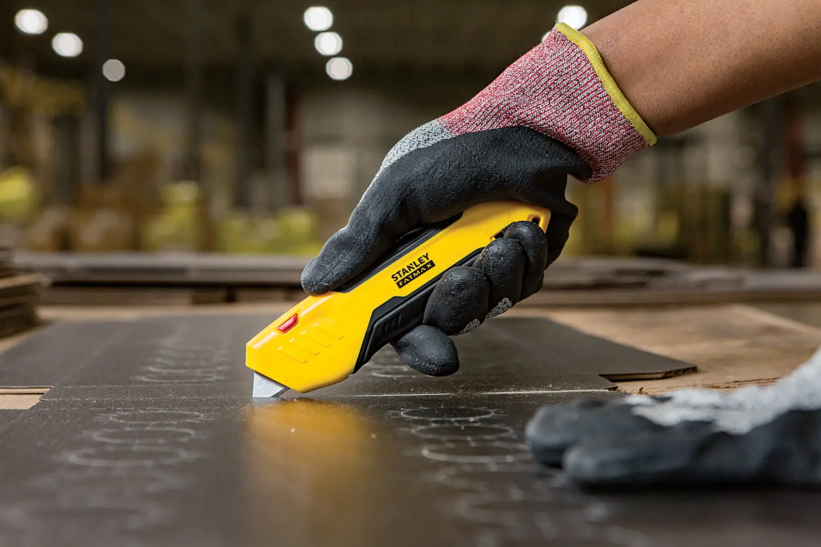 Stanley FMHT10369-0 utility knife cutting material, held by gloved hand in industrial setting.