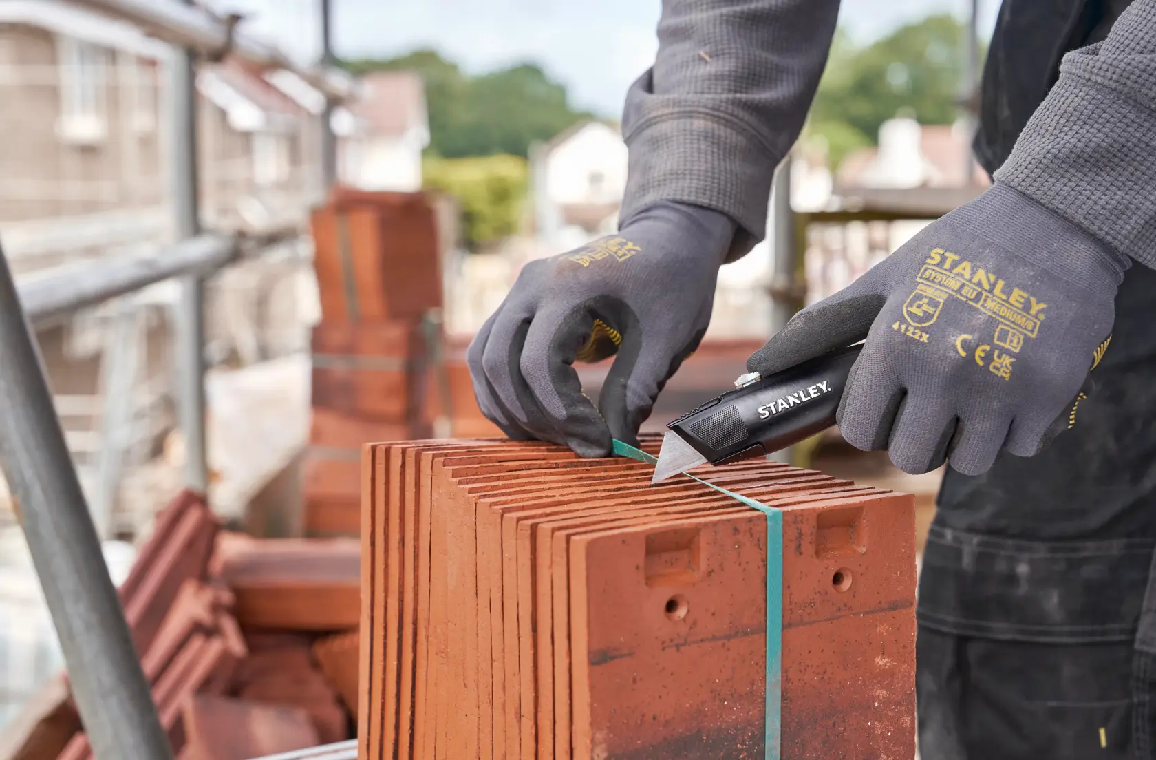 STANLEY FMHT10504-0 utility knife cutting green strap on stacked red tiles at construction site, close-up.