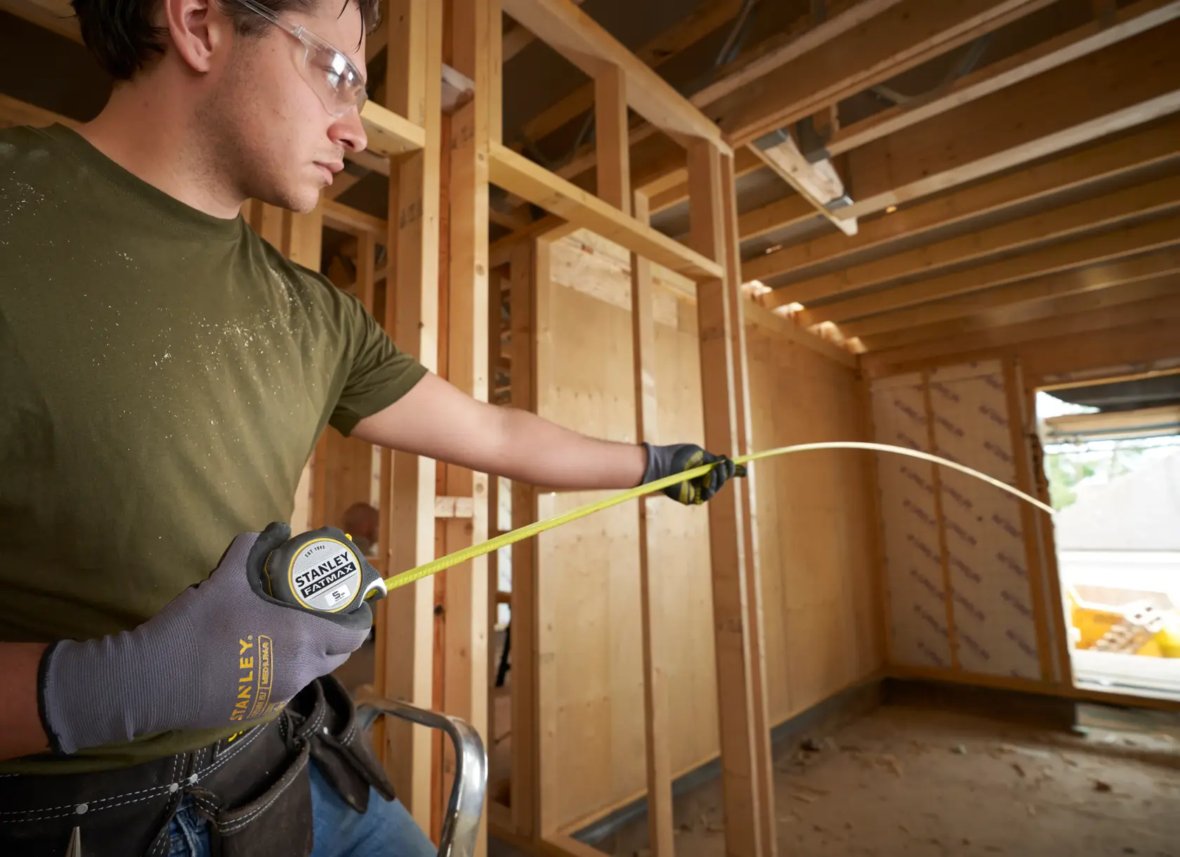 Stanley FMHT38214-0 tape measure used by worker in gloves at wood frame construction site.
