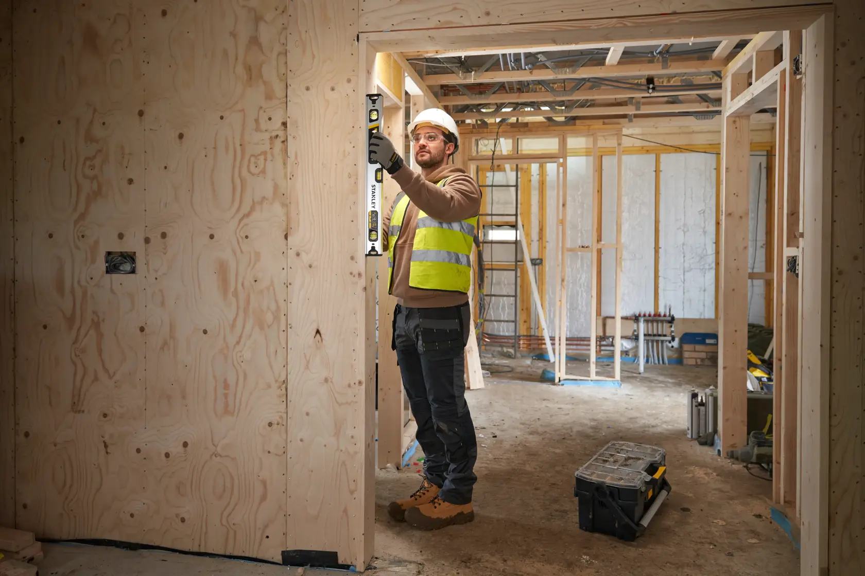 Worker uses STANLEY FMHT43672 spirit level on wooden door frame at construction site, wearing safety vest and boots.