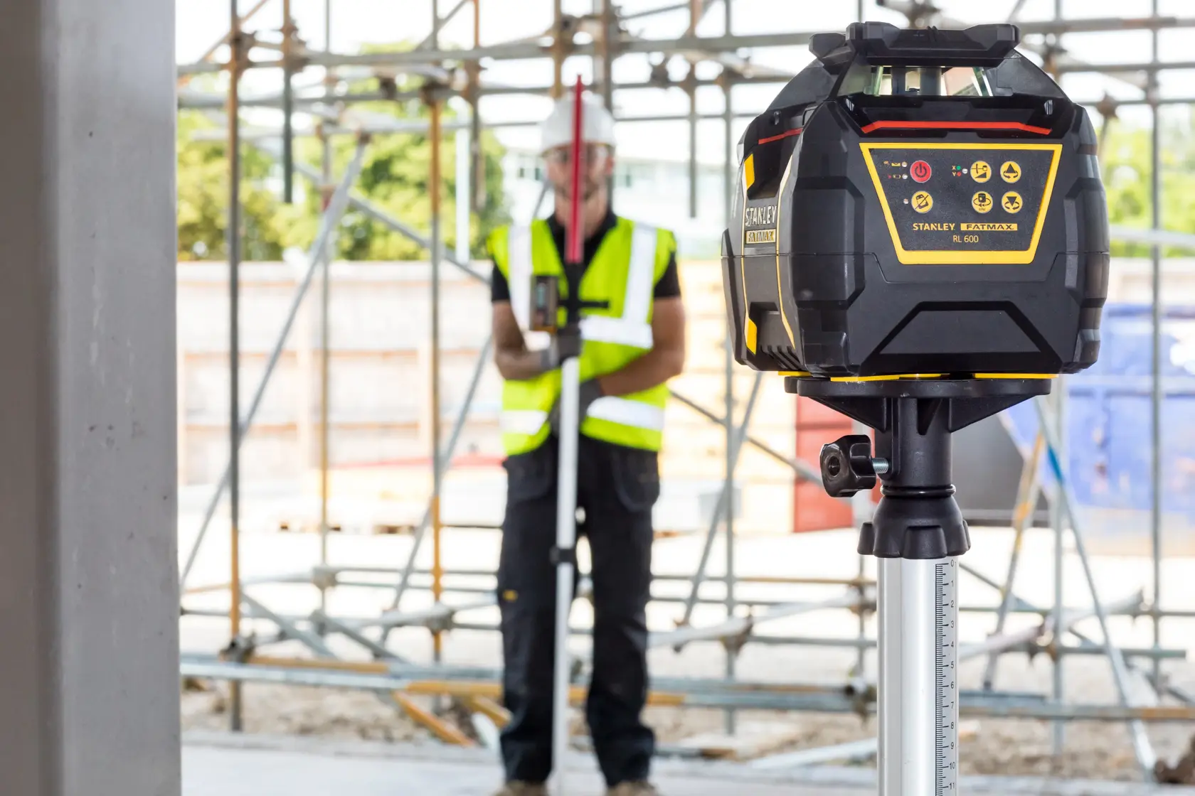 STANLEY FMHT77446-1 rotary laser level on tripod at construction site, worker in background.