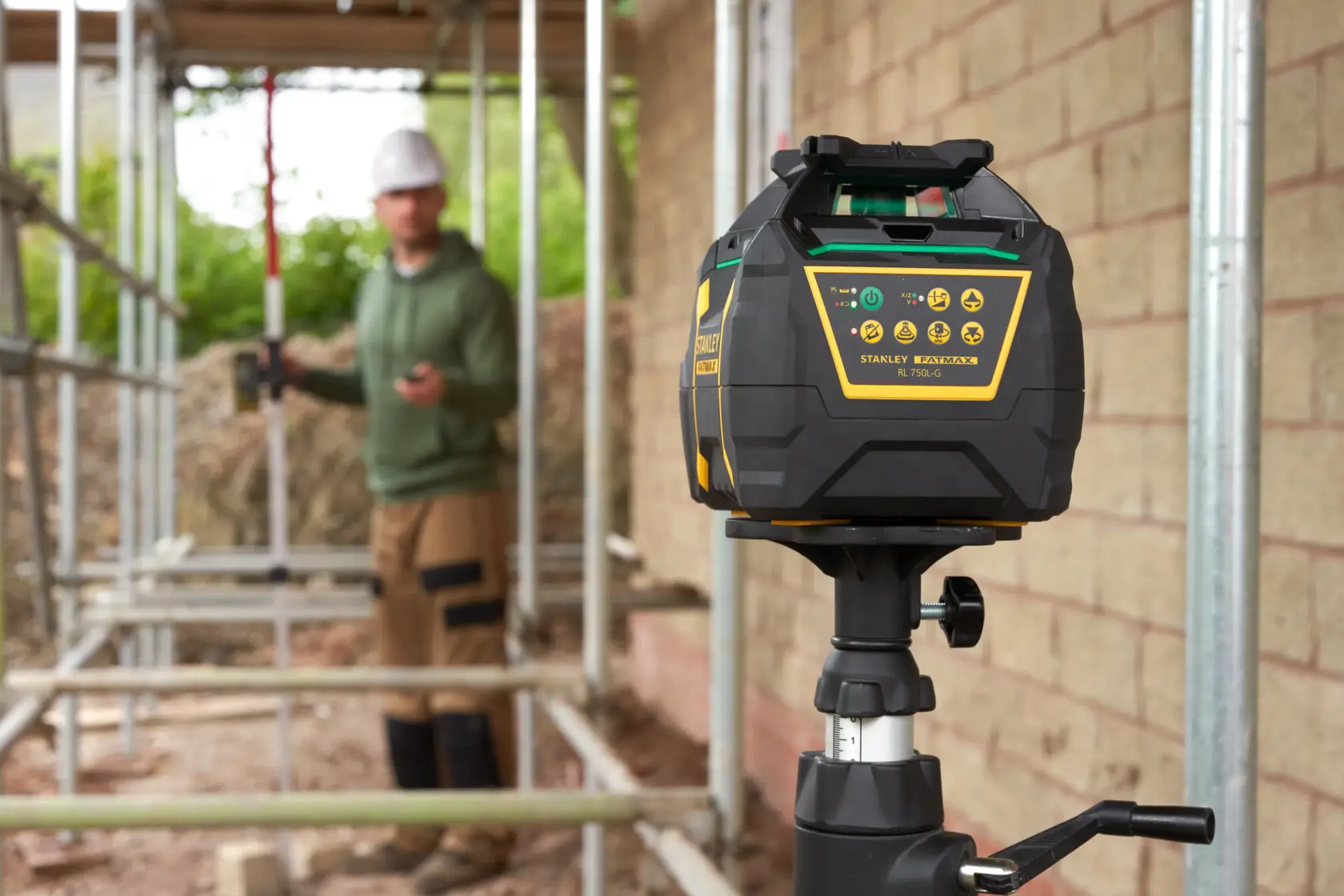 Photo of STANLEY FMHT77448 laser level on tripod at construction site, control panel visible, blurred worker in background.