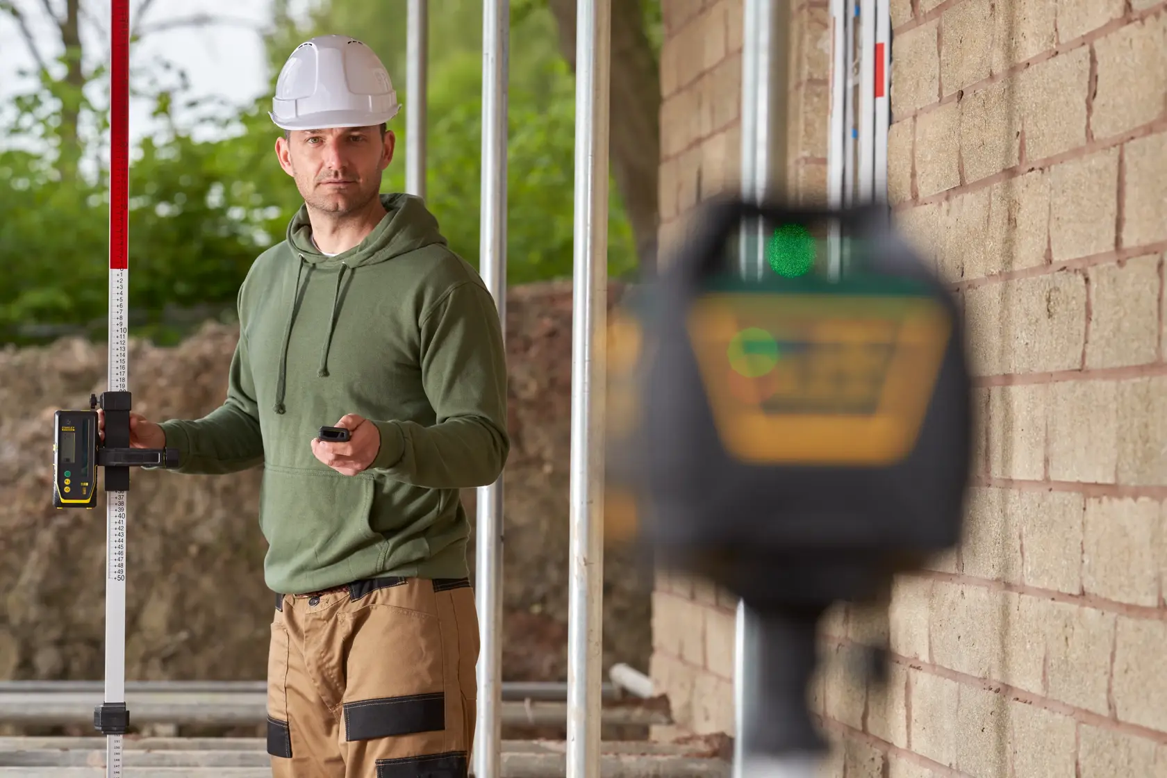 STANLEY FMHT77448 laser level in use at a construction site, worker holding measuring rod and remote control.