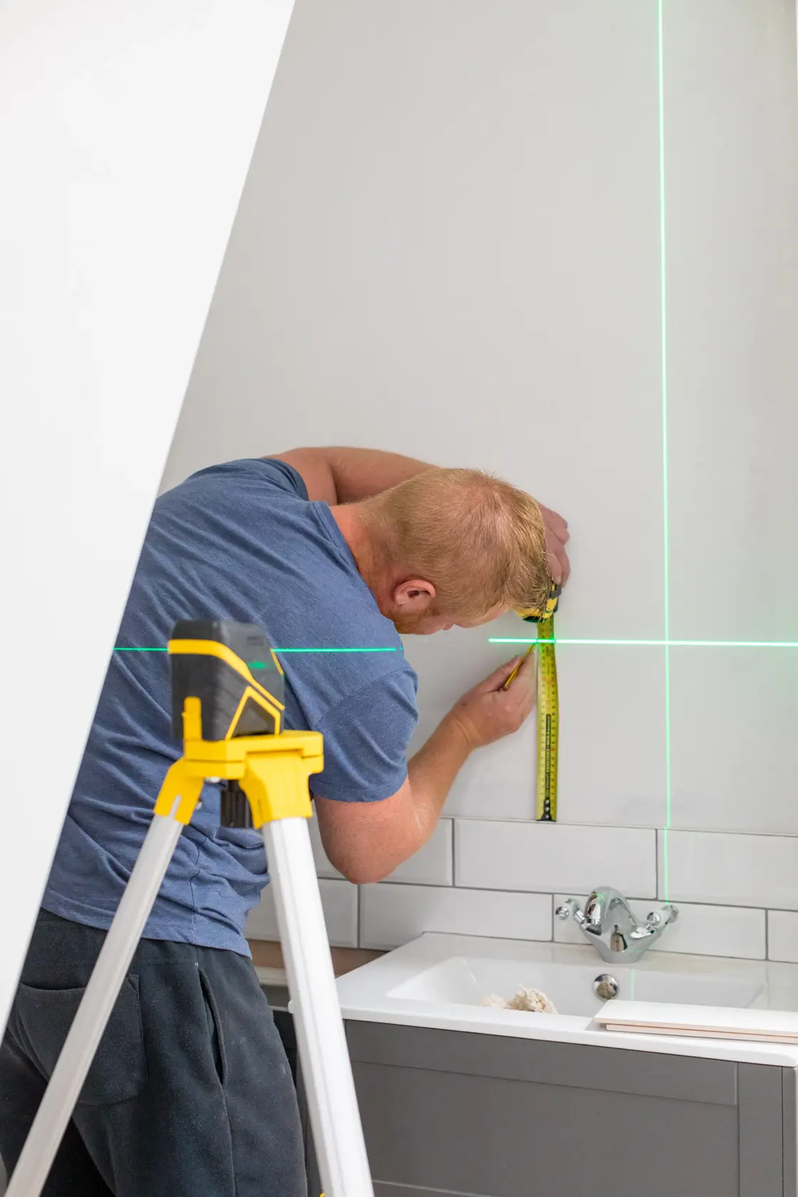 STANLEY FMHT77586-1 laser level on tripod projects green lines while man measures wall in bathroom.