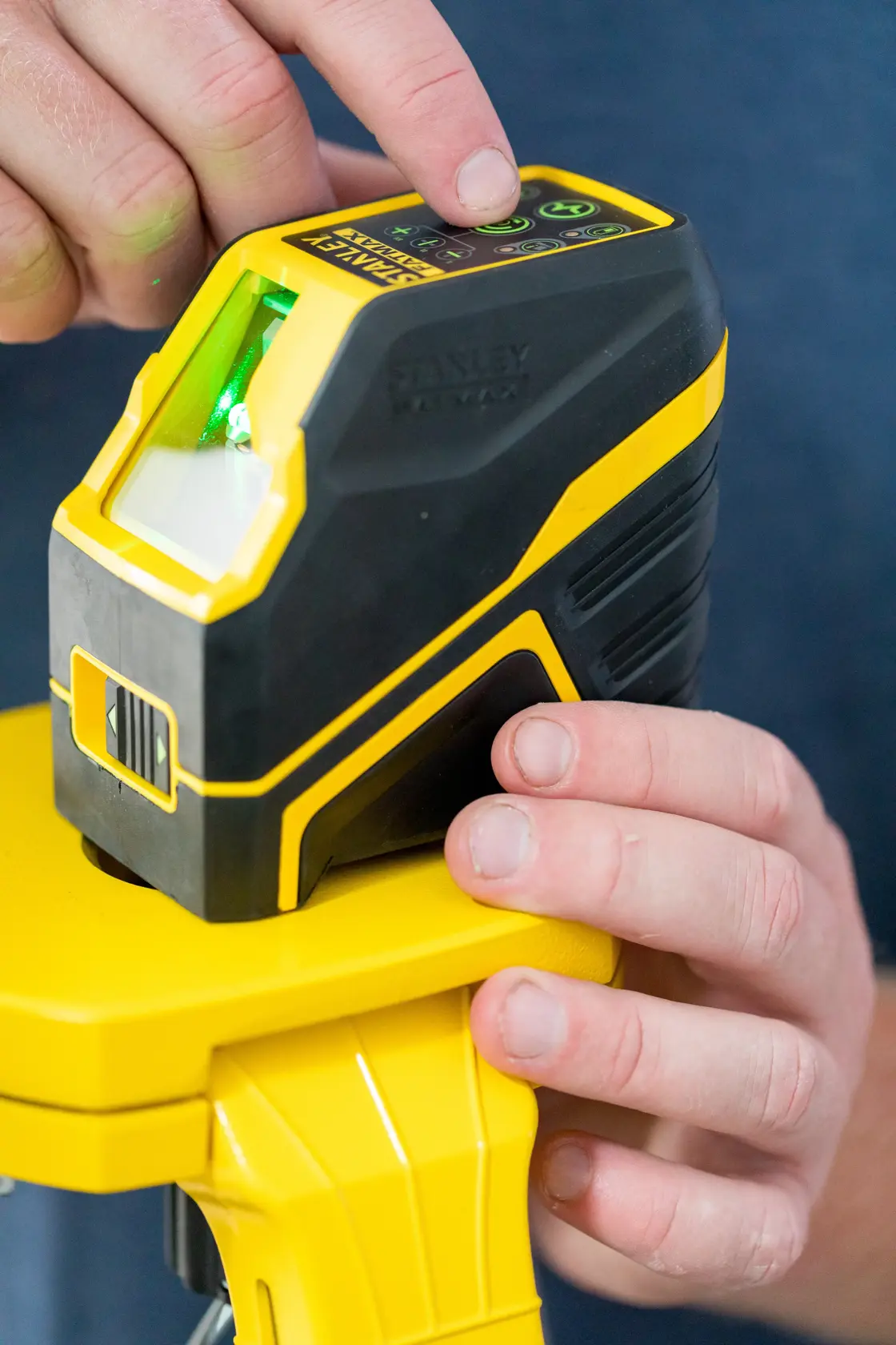 STANLEY FMHT77586-1 cross line laser level with green beam on tripod, close-up of hands operating top buttons.
