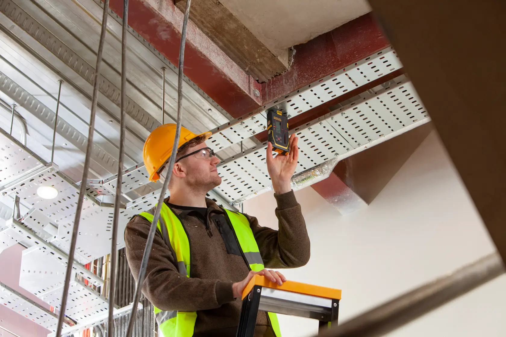 Photography of STANLEY FMHT77652-0 digital detector used by worker in safety gear on ladder at construction site.