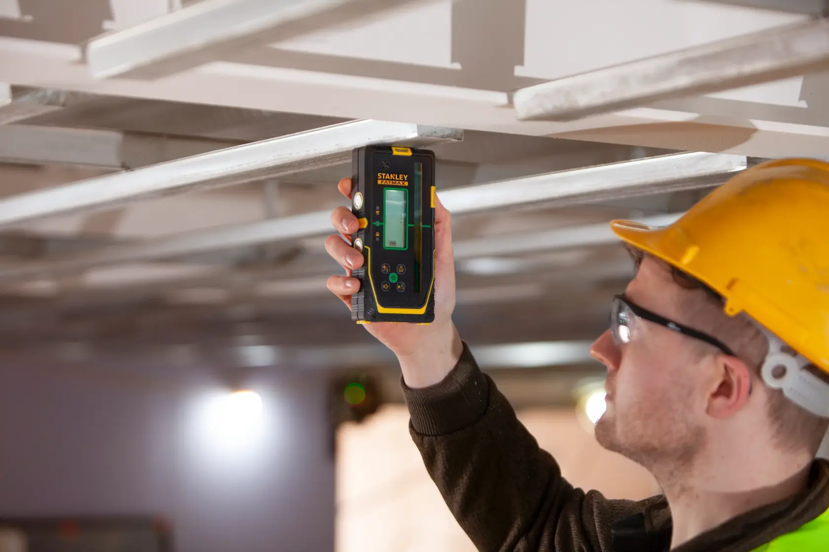 STANLEY FMHT77653-0 laser level held by worker in yellow helmet measuring metal ceiling frame.