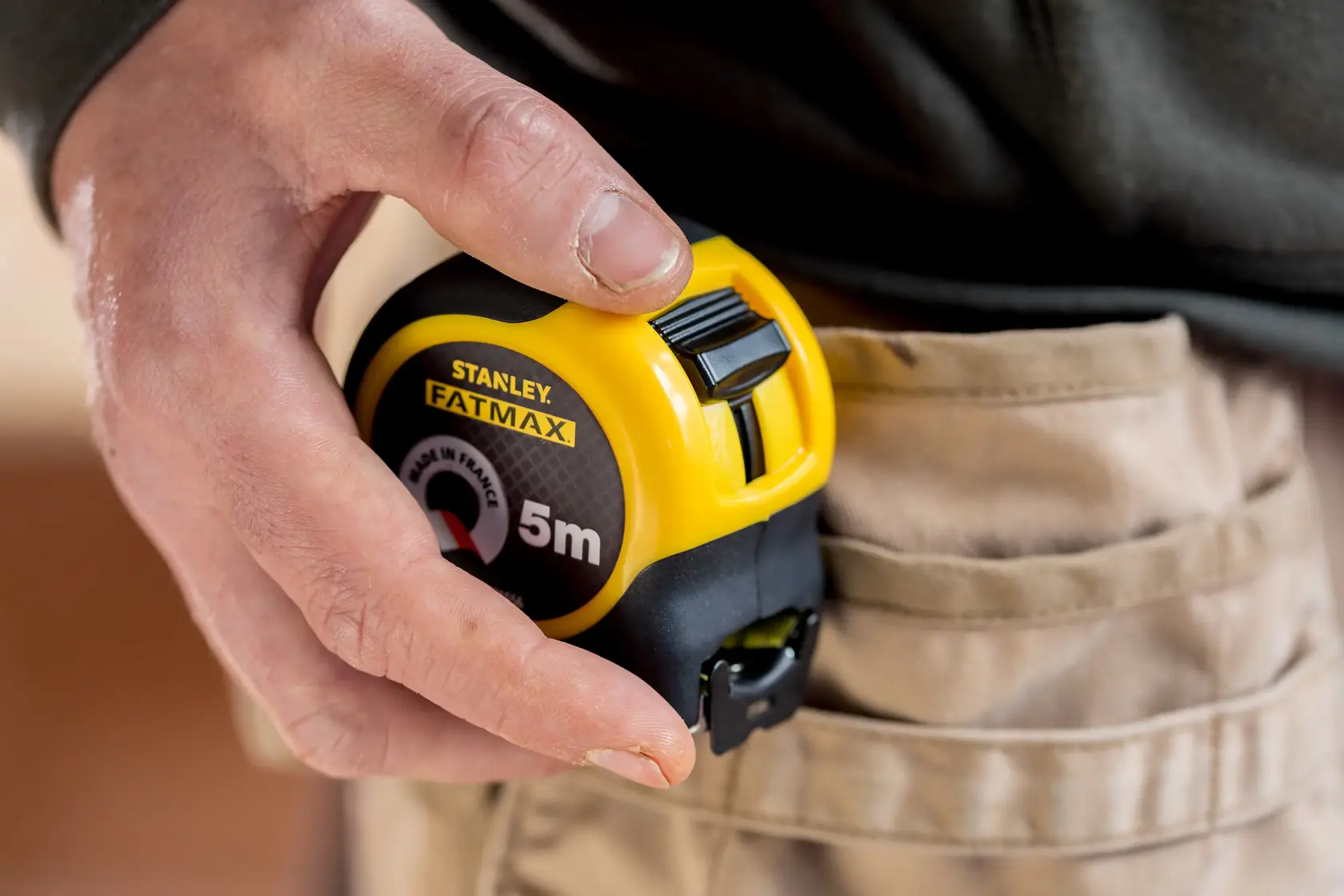 STANLEY FMHT81556-0 tape measure, yellow and black, held by hand next to beige work trousers.
