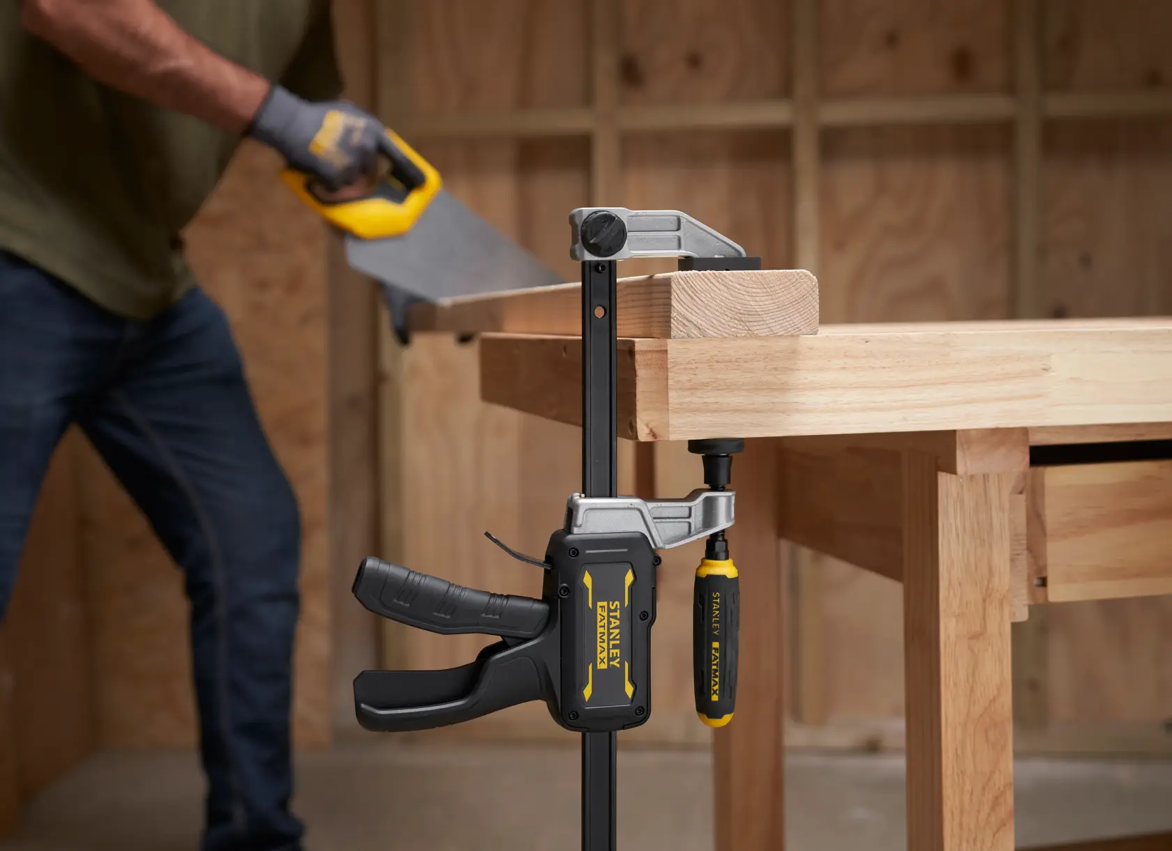 STANLEY FMHT83007-0 clamp securing wood to workbench, close-up, with person sawing in background.