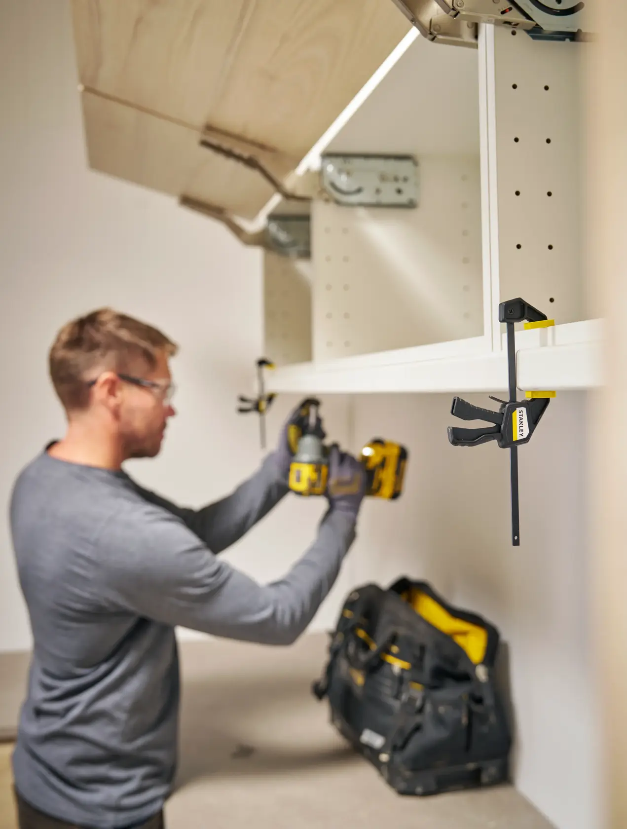 Photo of a STANLEY FMHT83270-0 bar clamp holding a white shelf, with a tool bag and person using a drill nearby.