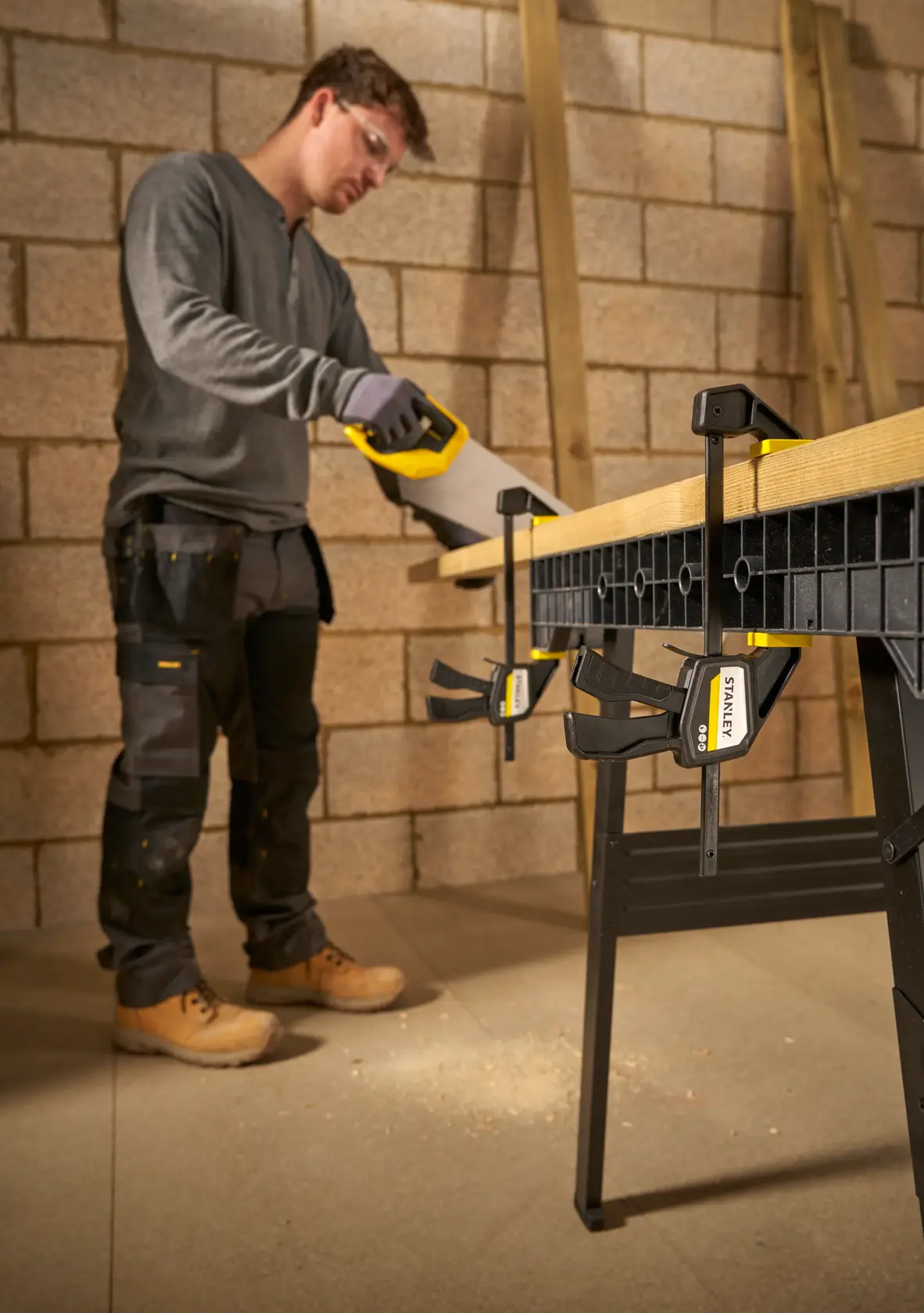 Photo of STANLEY FMHT83271-0 clamp securing wood to workbench in a workshop, worker sawing near clamps.