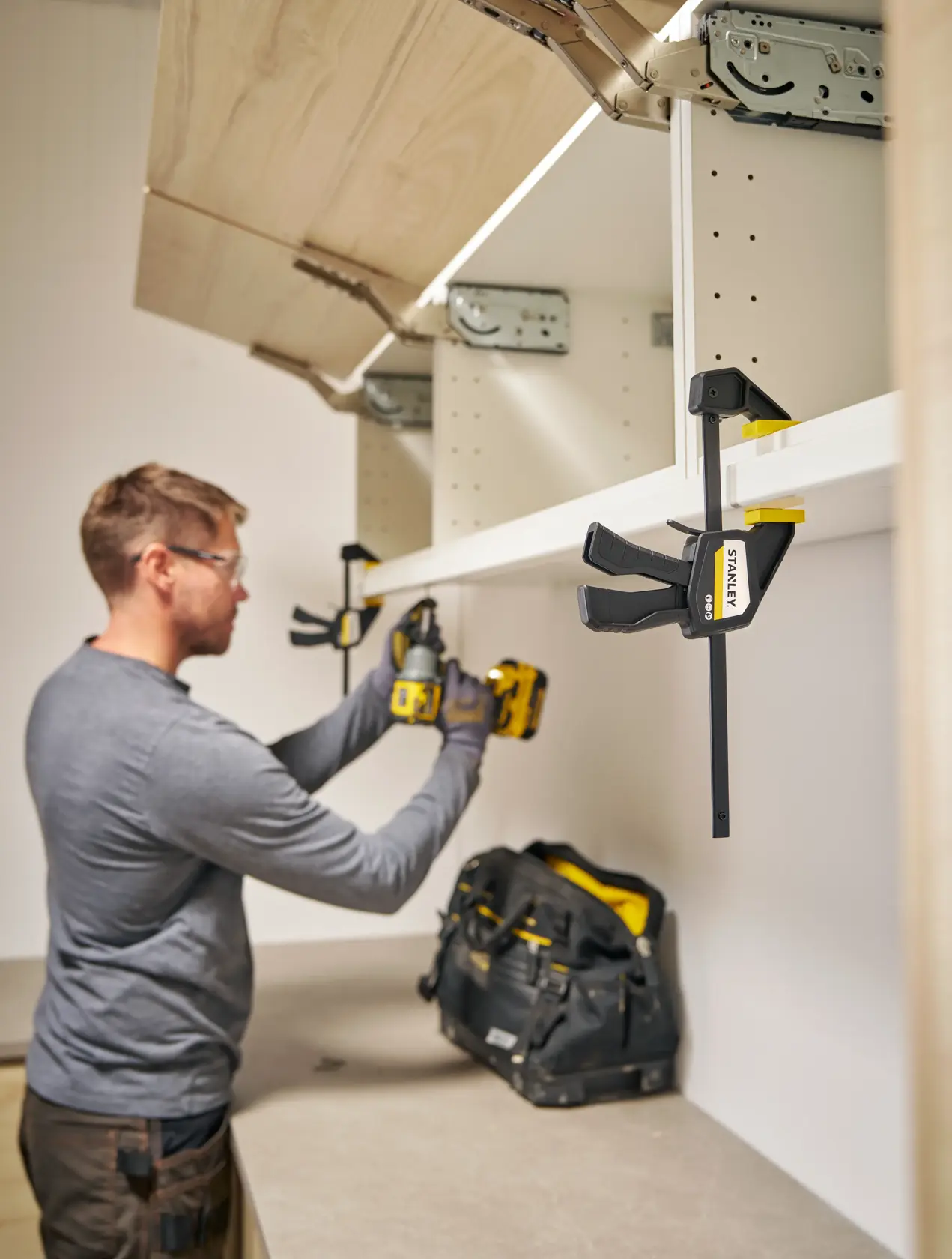 Photo of STANLEY FMHT83271-0 clamp holding a shelf with tool bag and person assembling cabinet.