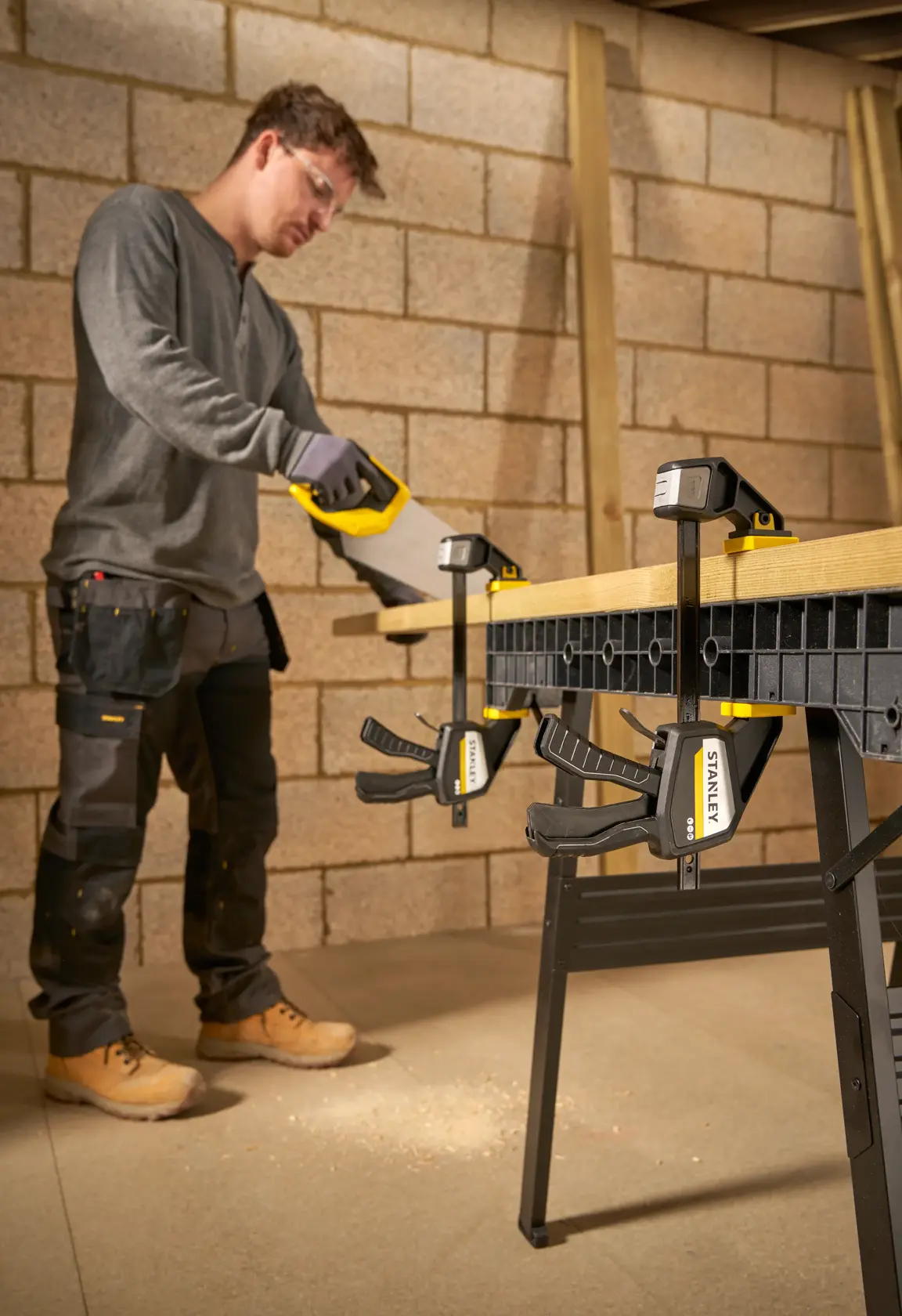 STANLEY FMHT83273-0 clamp securing wood to workbench, person sawing in workshop setting.