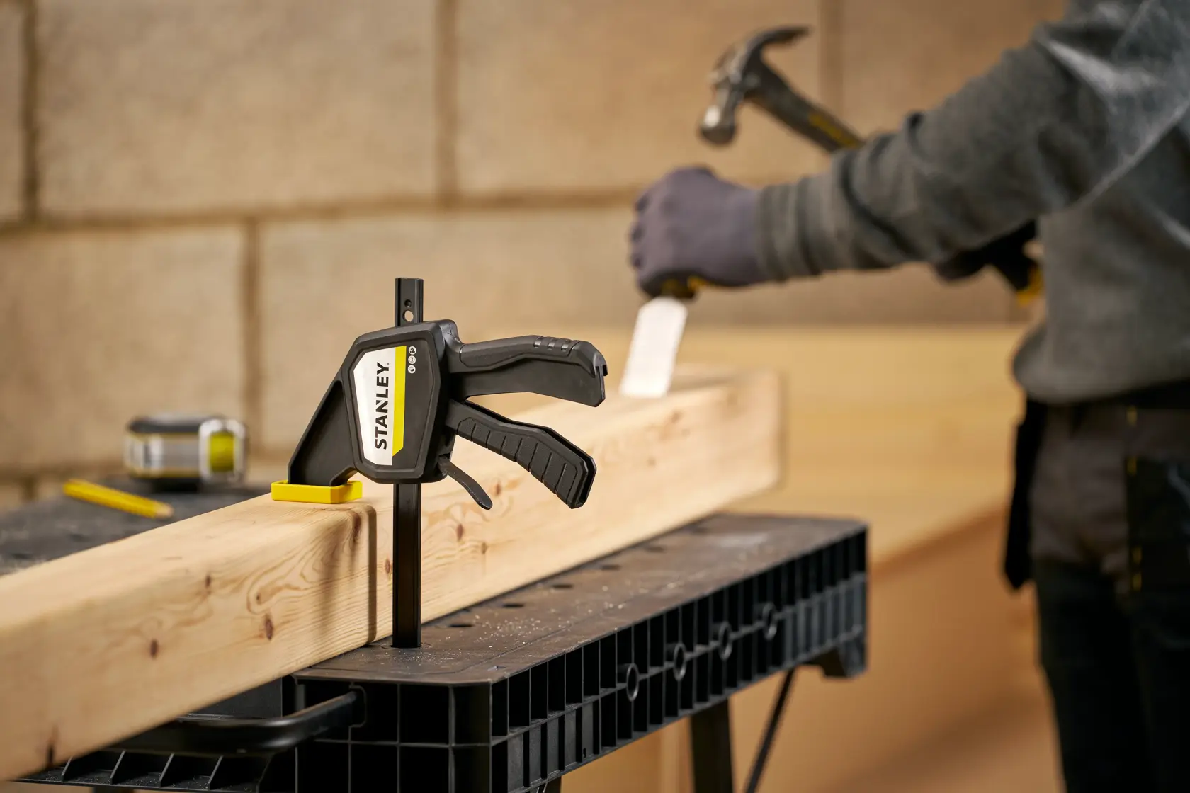 STANLEY FMHT83273-0 clamp securing wood beam on workbench with worker using hammer in background.