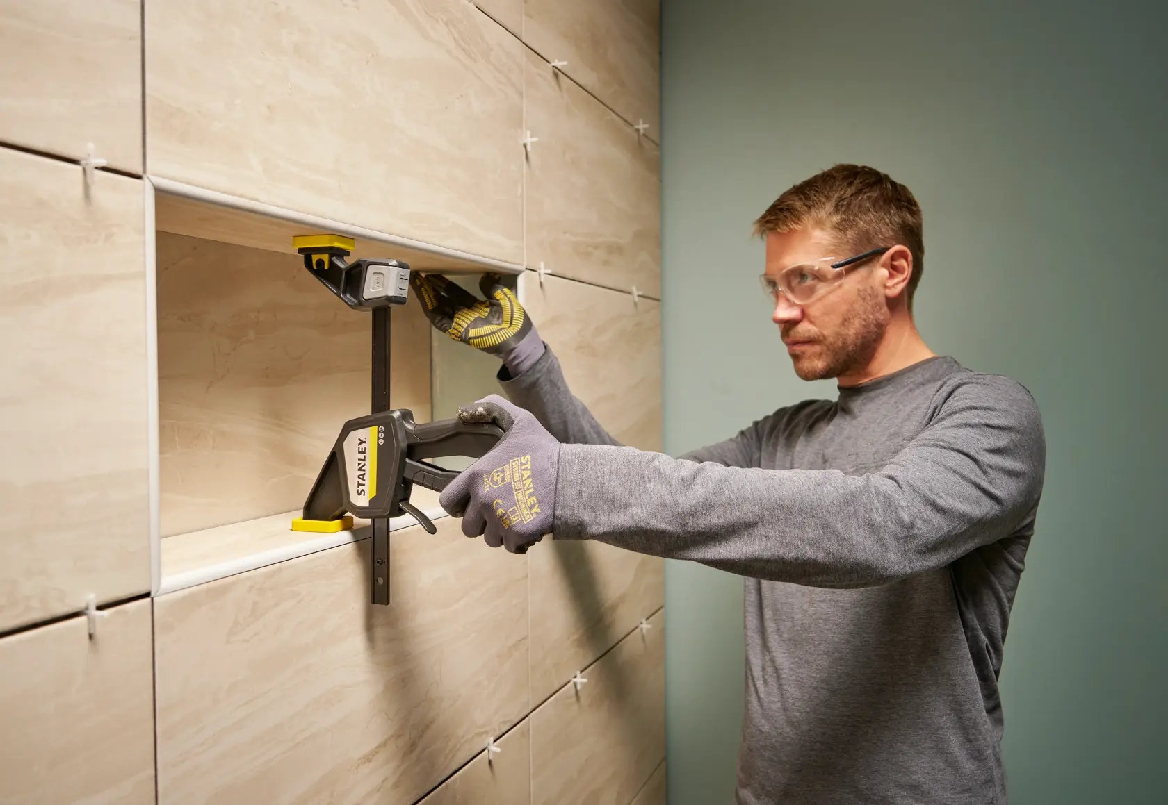 STANLEY FMHT83273-0 clamp in use on a tiled wall, person wearing gloves holding the clamp.