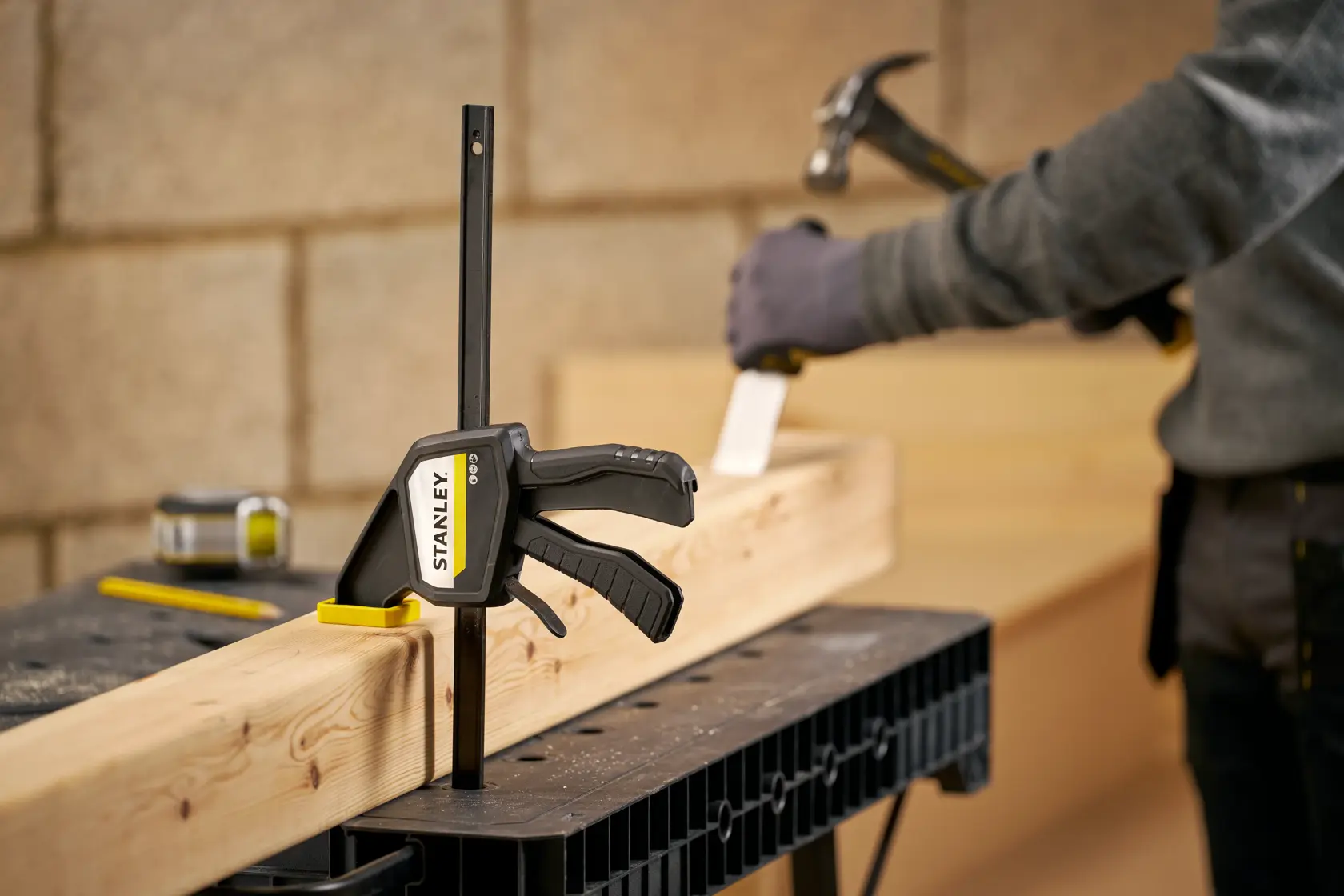 STANLEY FMHT83274-0 bar clamp secures wood beam on workbench, person using hammer in background.