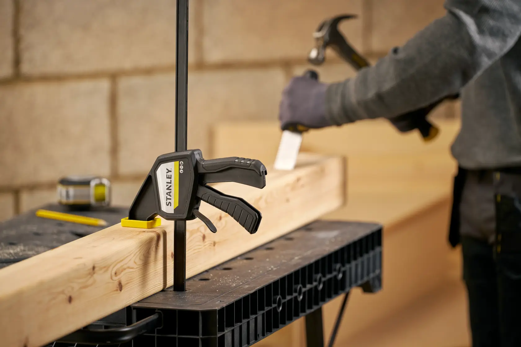 STANLEY FMHT83275-0 bar clamp securing wood beam on workbench, person using hammer in background.