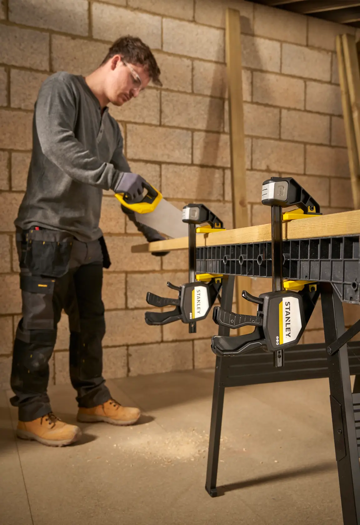 Photo of STANLEY FMHT83278-0 clamps holding wood on a workbench while a person saws, workshop background.