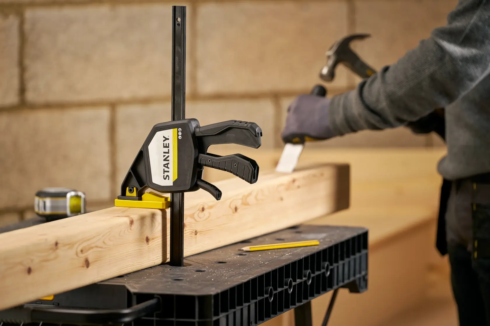 STANLEY FMHT83279-0 bar clamp securing wood on workbench, worker with hammer in background, workshop setting.