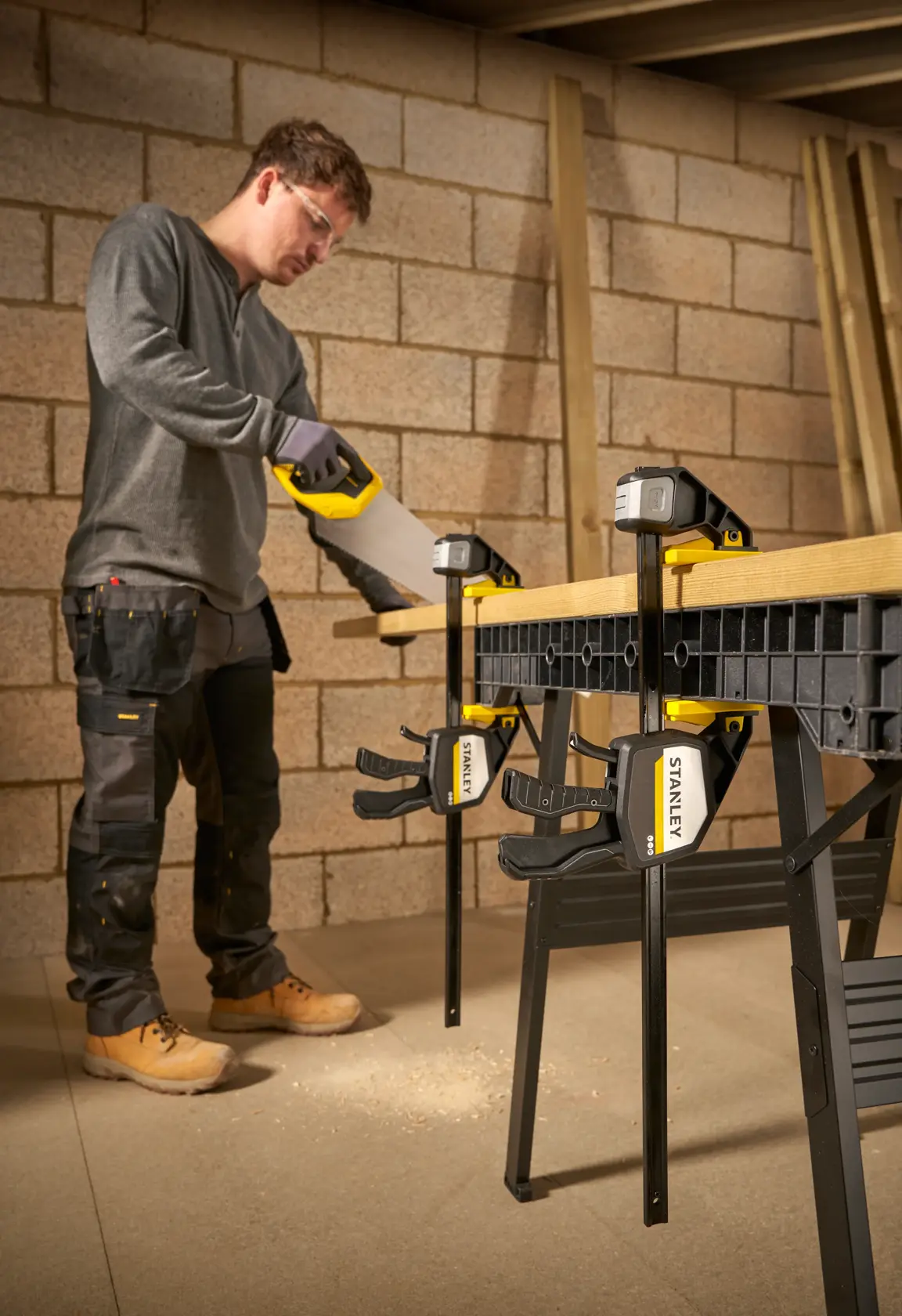 STANLEY FMHT83280-0 clamp securing wood to workbench, person sawing in workshop setting.