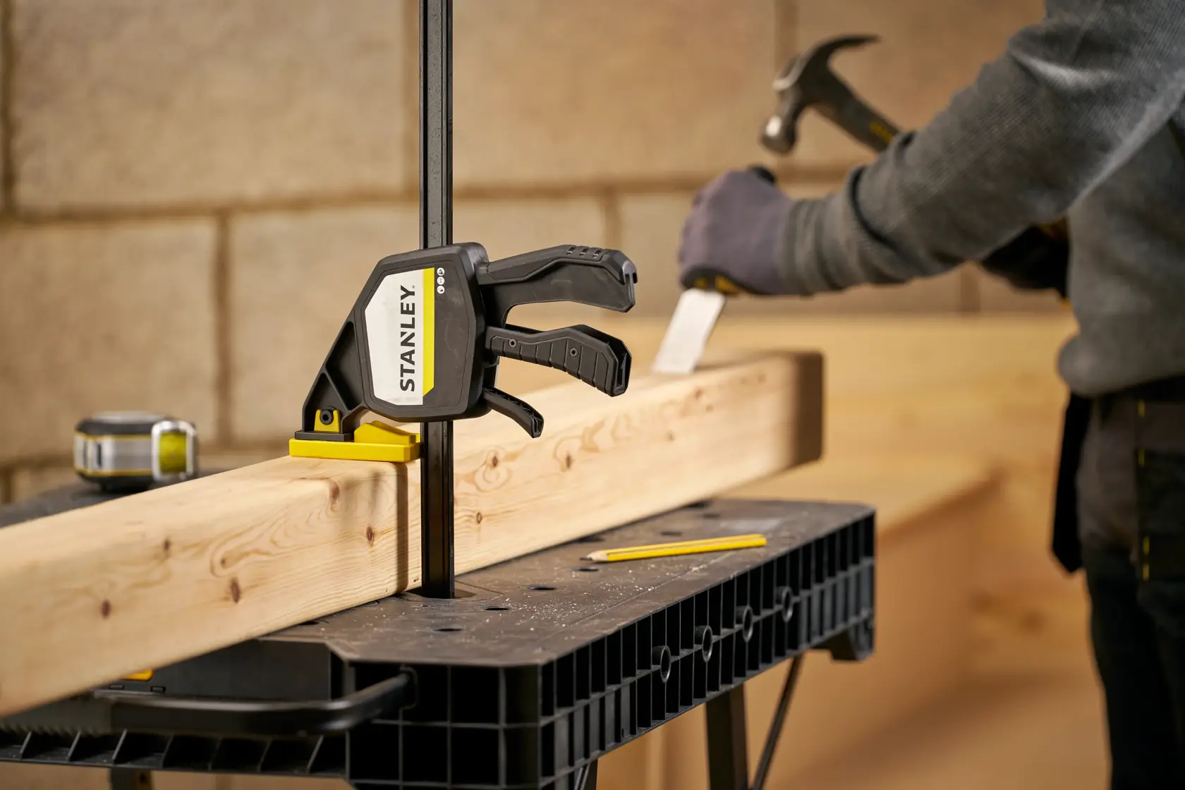 Photo of STANLEY FMHT83280-0 trigger clamp securing wood on workbench with person working in background.