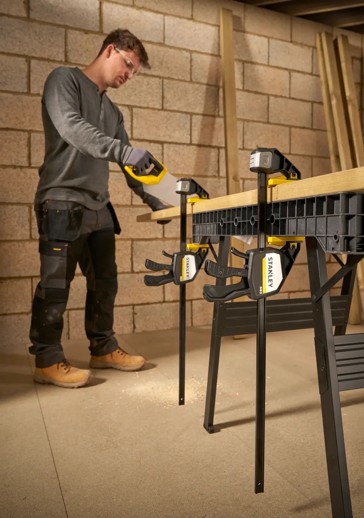 STANLEY FMHT83281-0 clamp securing wood on workbench with person sawing, workshop setting.