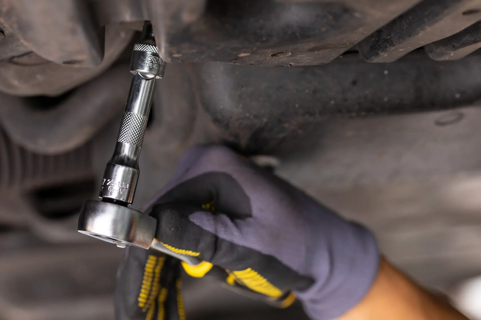 STANLEY FMMT25130-0 ratchet being used under a vehicle, close-up of gloved hand operating tool.