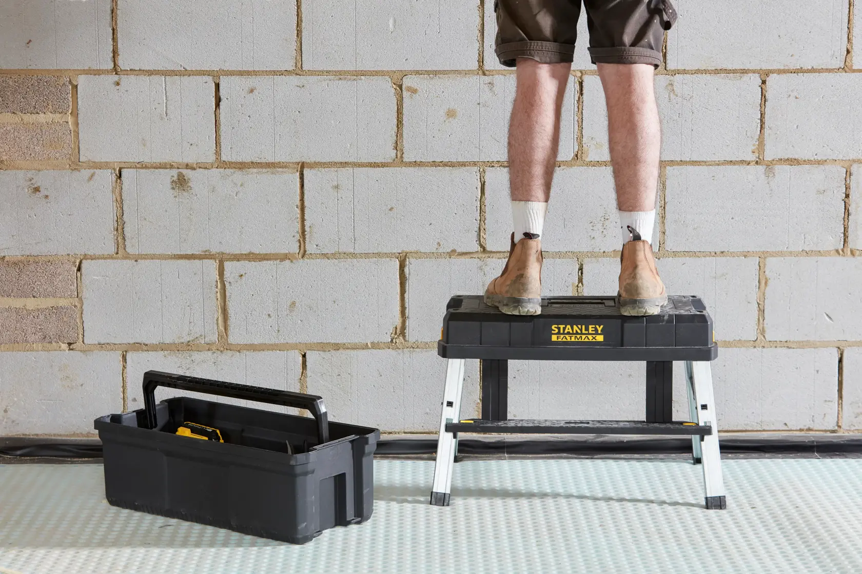 STANLEY FMST81083-1 step stool with man standing, black tool tote on floor, indoor setting.