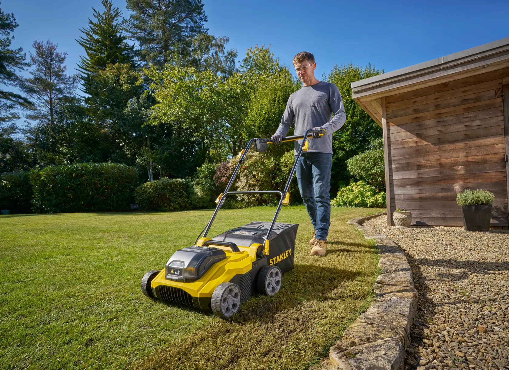 Photo of STANLEY SCOEP3801M2 lawn scarifier in use on grass near a wooden shed, with person pushing the machine outdoors.