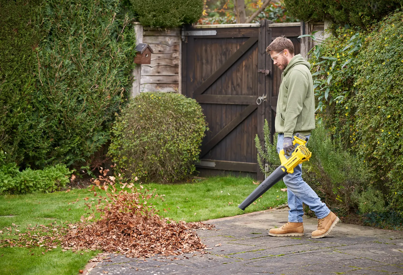 STANLEY SFMCBL714B-XJ leaf blower in use, blowing leaves on a garden path, side outdoor view.