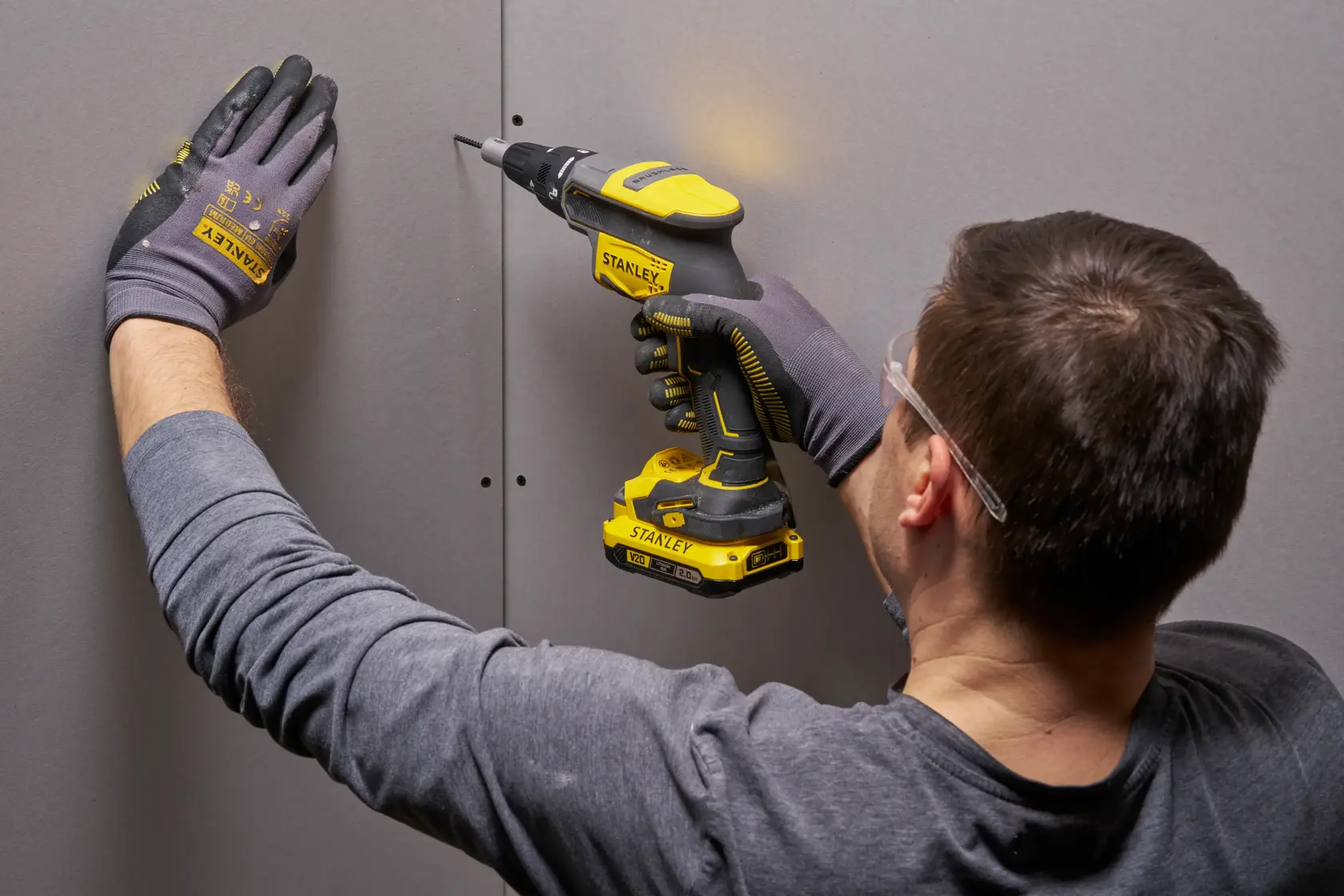 Photo of worker using STANLEY SFMCF600D1S cordless screwdriver on drywall, yellow and black tool, side angle.