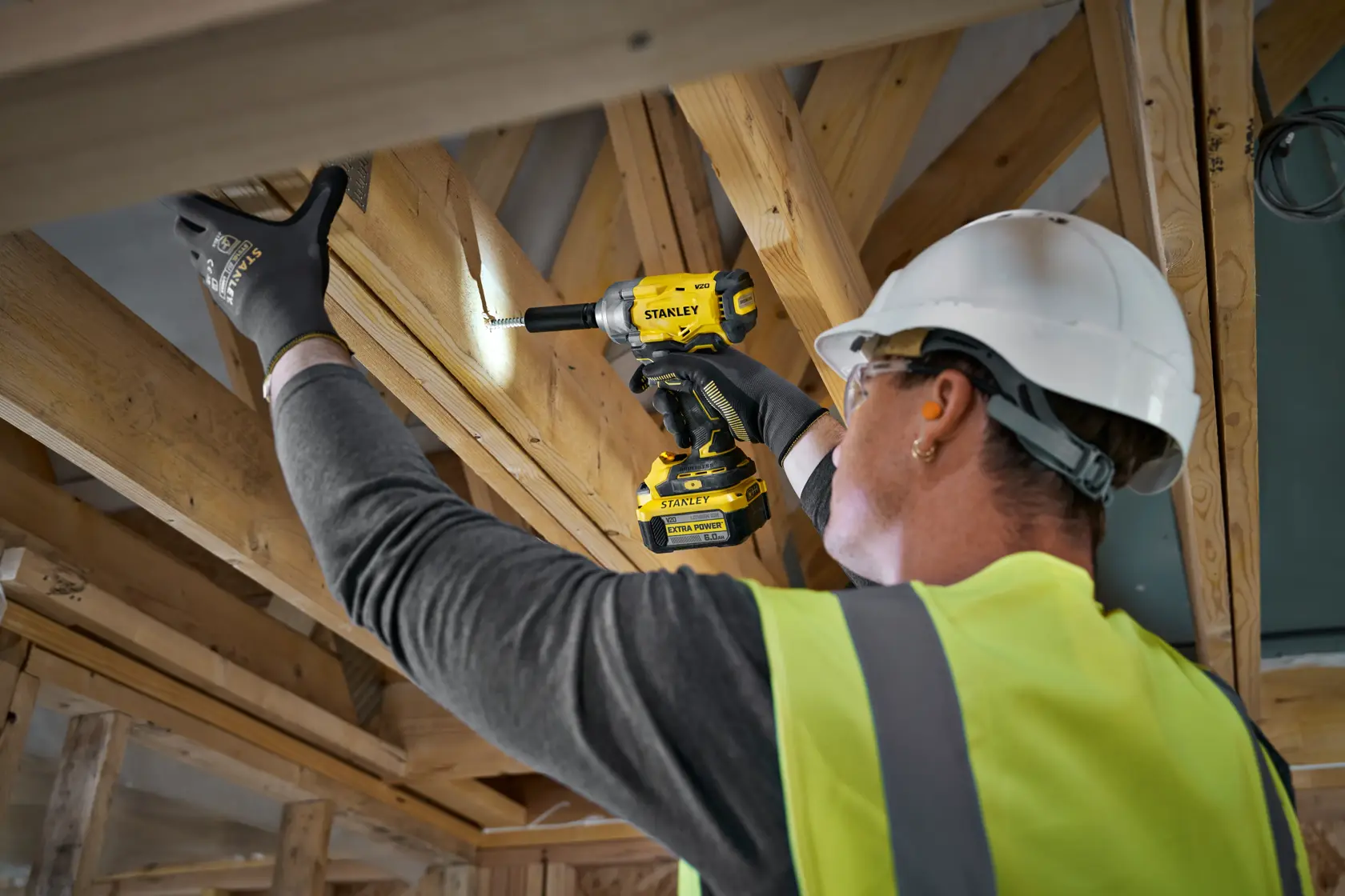 Worker using a STANLEY cordless drill on wooden beams, wearing gloves, helmet, and yellow vest at construction site.