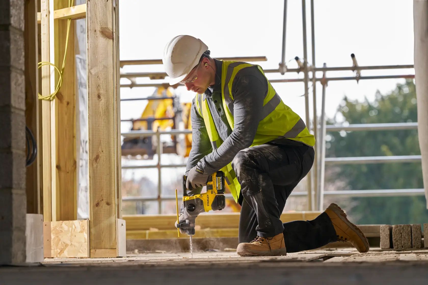 Worker in safety gear uses a yellow Stanley drill on concrete at a construction site.