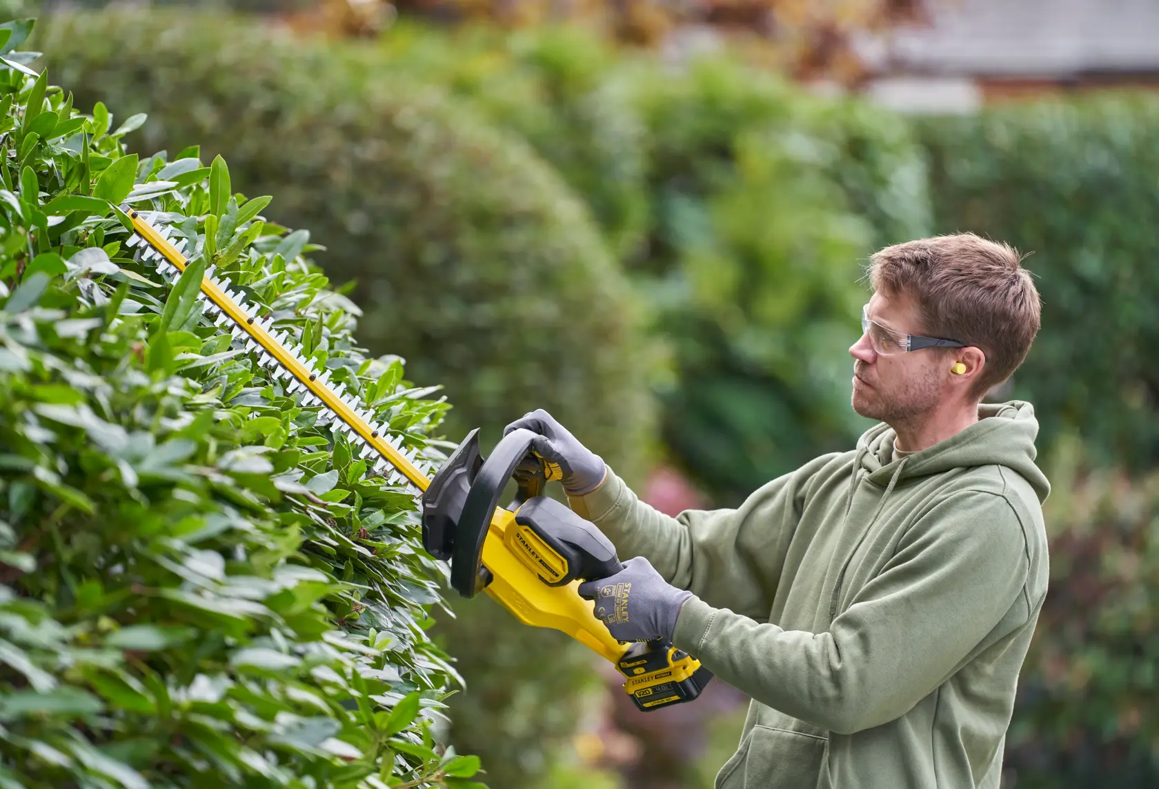 Photo of STANLEY SFMCHT655B hedge trimmer in use, yellow and black, trimming green bushes outdoors.