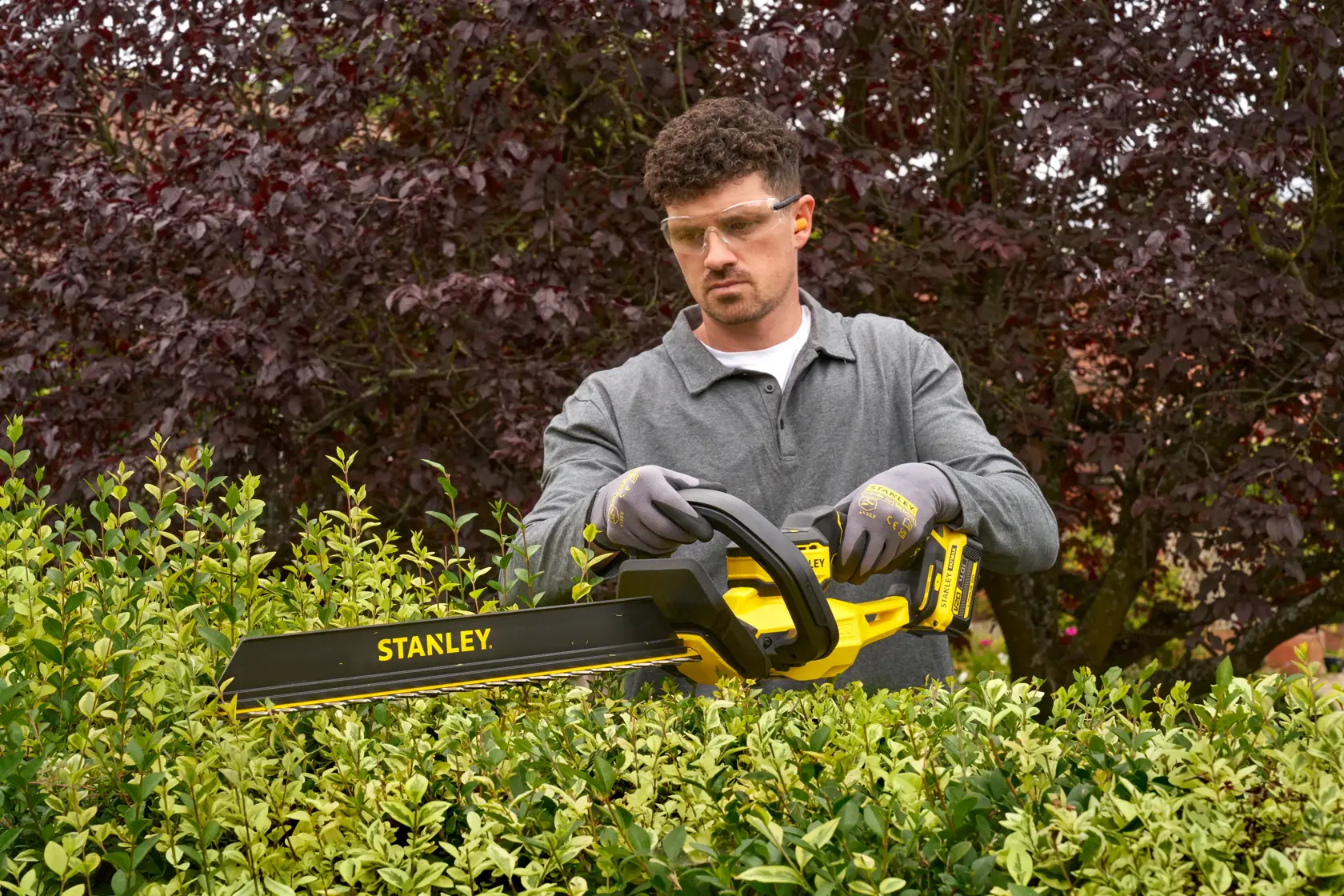 Photo of a person using a STANLEY STCHT8255B hedge trimmer in a garden, trimming green bushes.