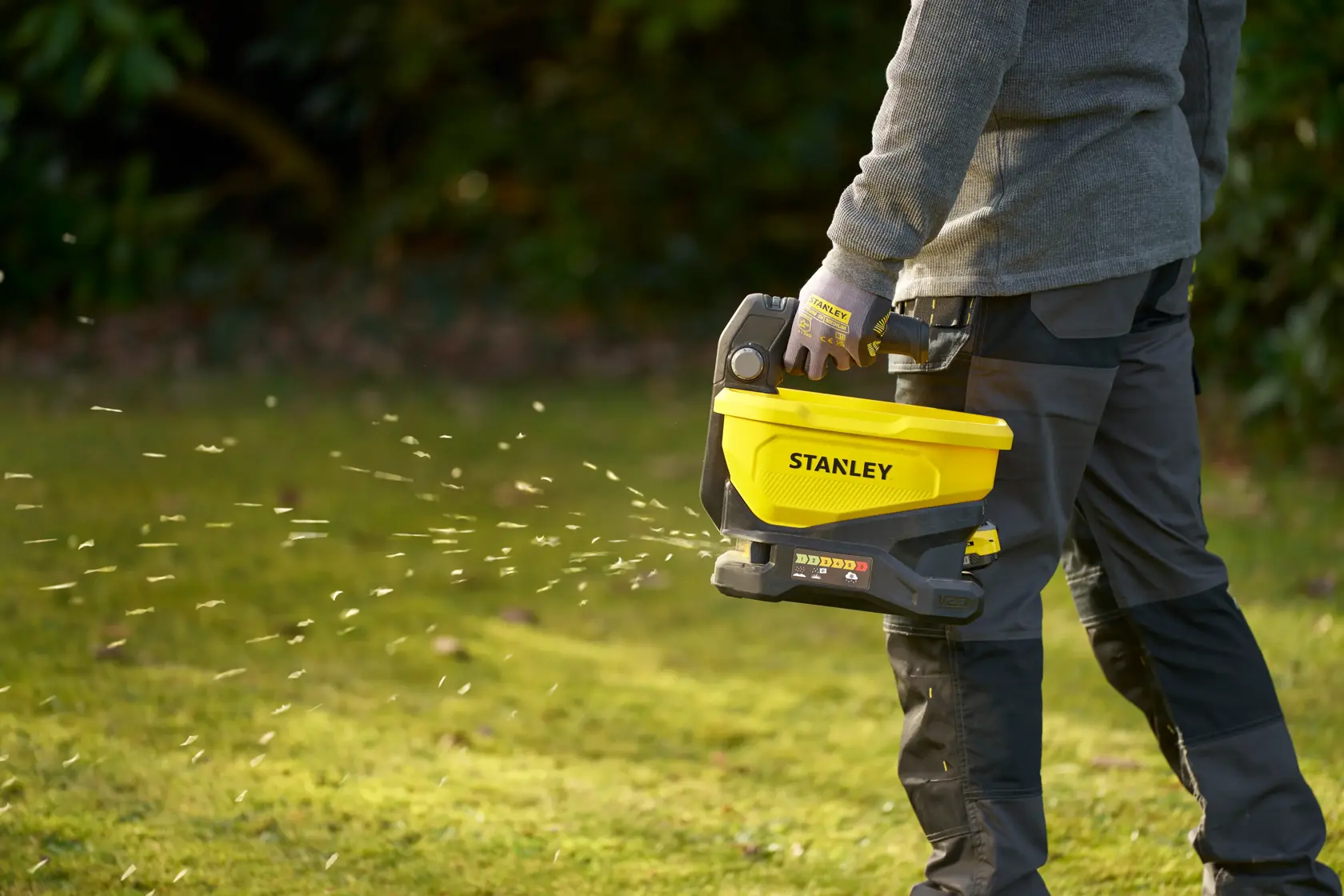 Photo of person using STANLEY STCOE300B handheld spreader in yellow and black, dispersing material on grass.