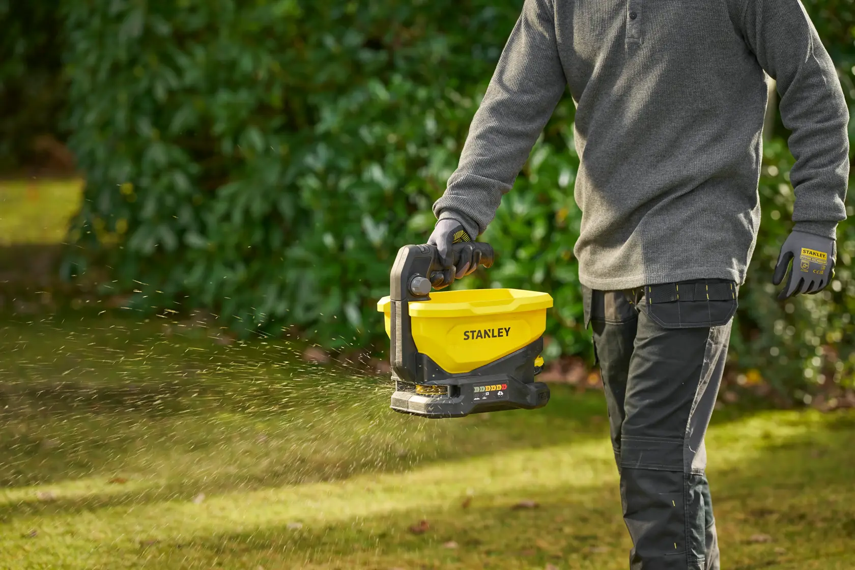 STANLEY STCOE300B spreader in use outdoors, yellow and black handheld tool dispersing material on grass.