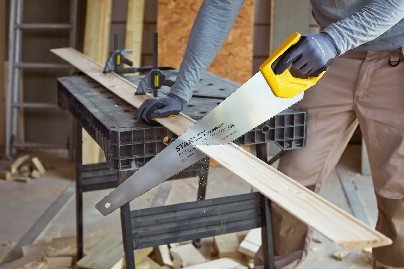 STANLEY STHT1-20353 hand saw with yellow handle cutting wood plank in workshop setting.