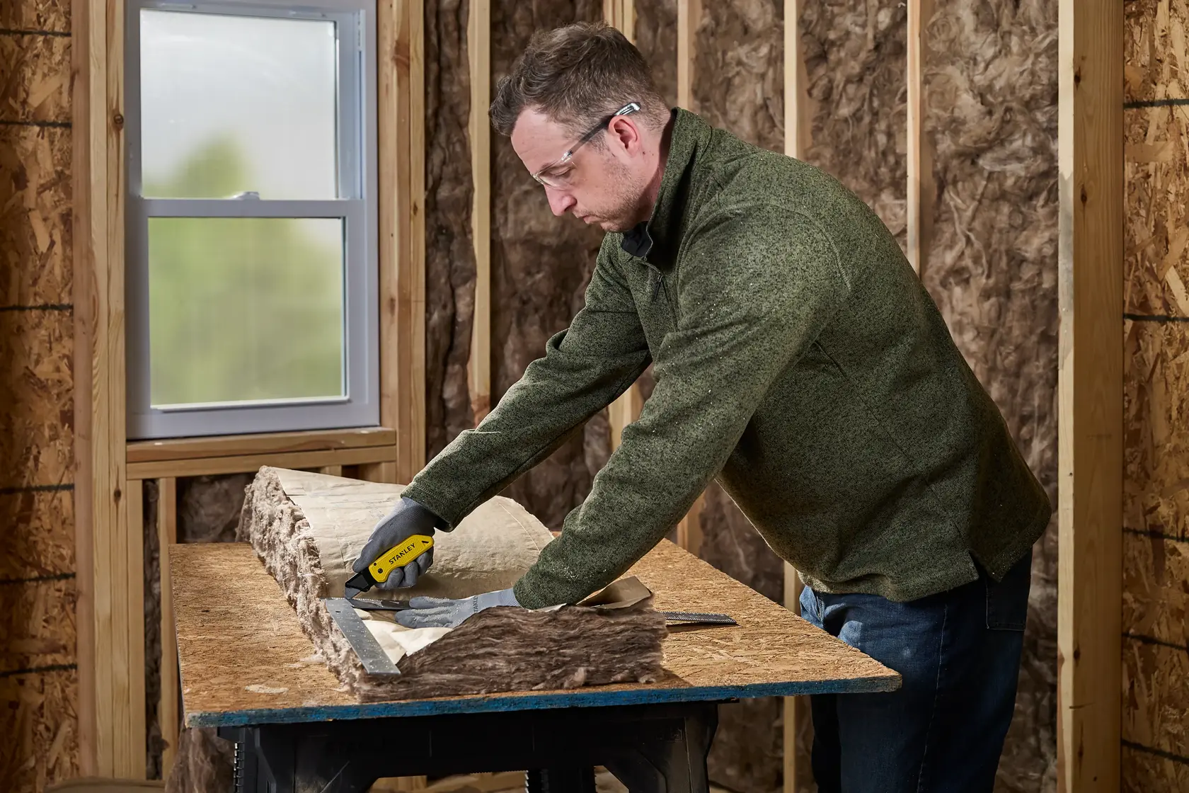 A person wearing gloves uses a yellow and black STANLEY utility knife to cut insulation material on a workbench in a room under construction.